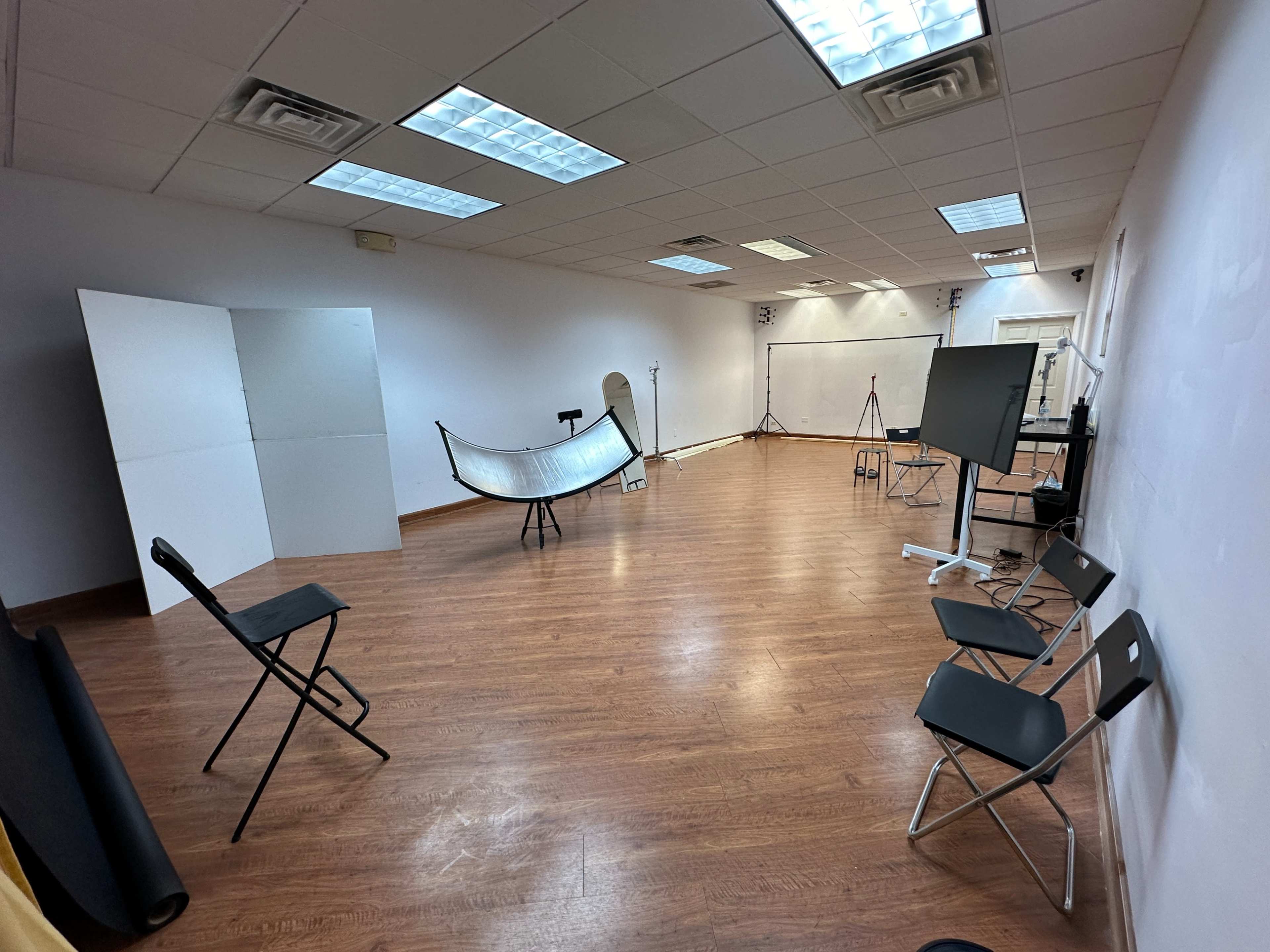 A spacious studio with wooden flooring, several chairs, and photography equipment arranged against the plain white walls.