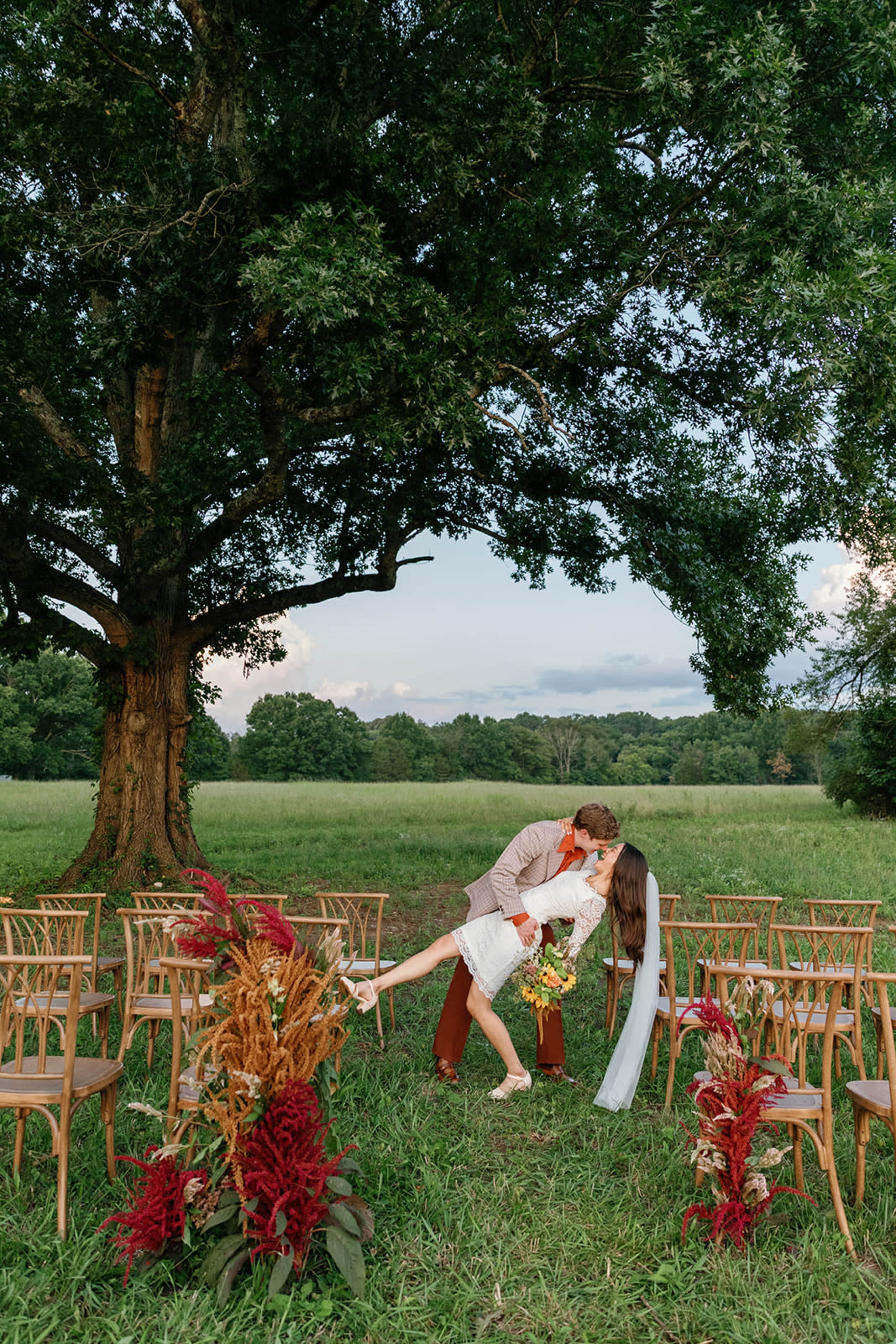 A couple shares a kiss in front of a wedding ceremony setup under a large tree, surrounded by wooden chairs and colorful floral arrangements.