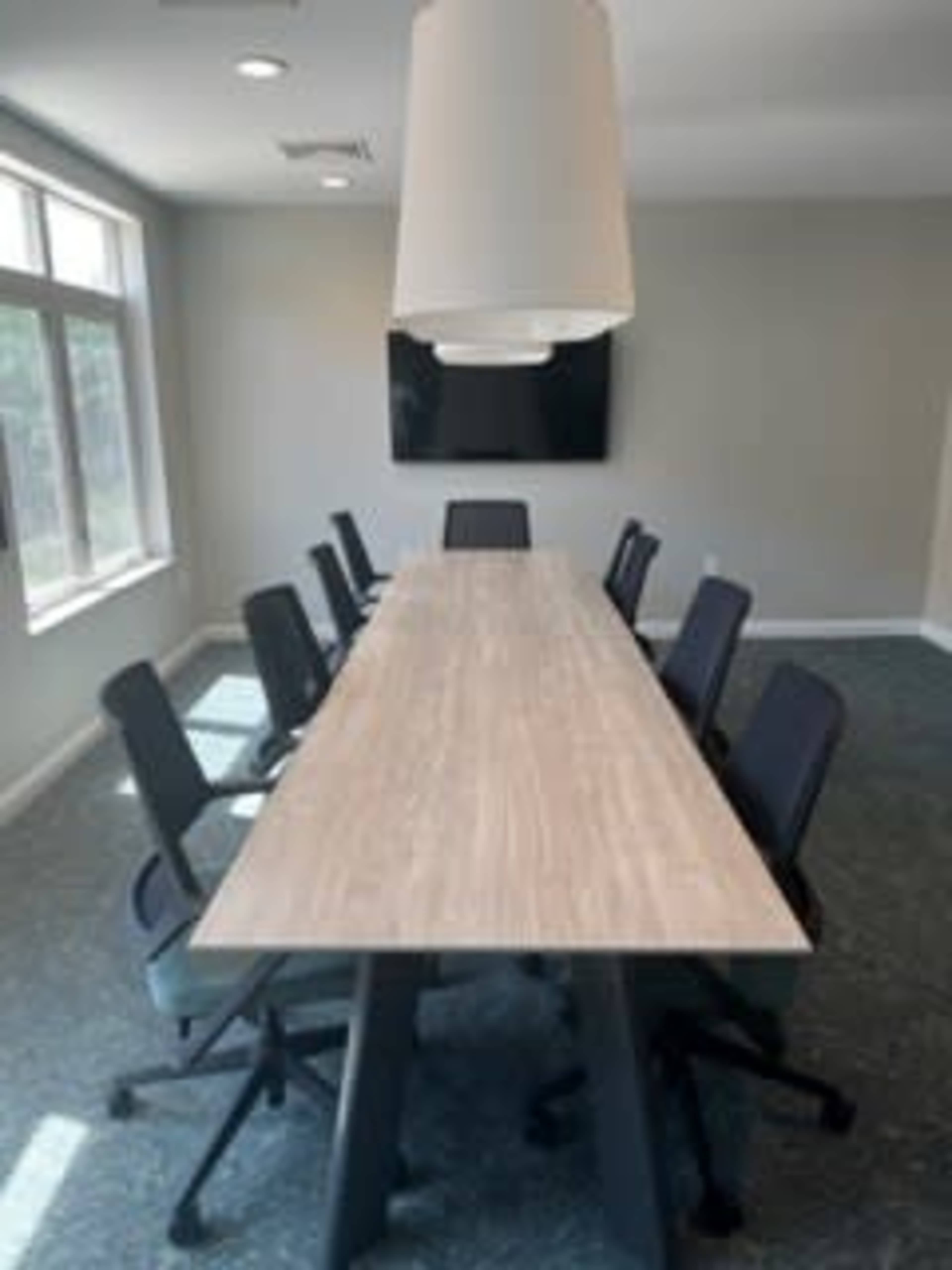 A long conference table with eight ergonomic chairs is set in a well-lit meeting room featuring large windows and a television mounted on the wall.
