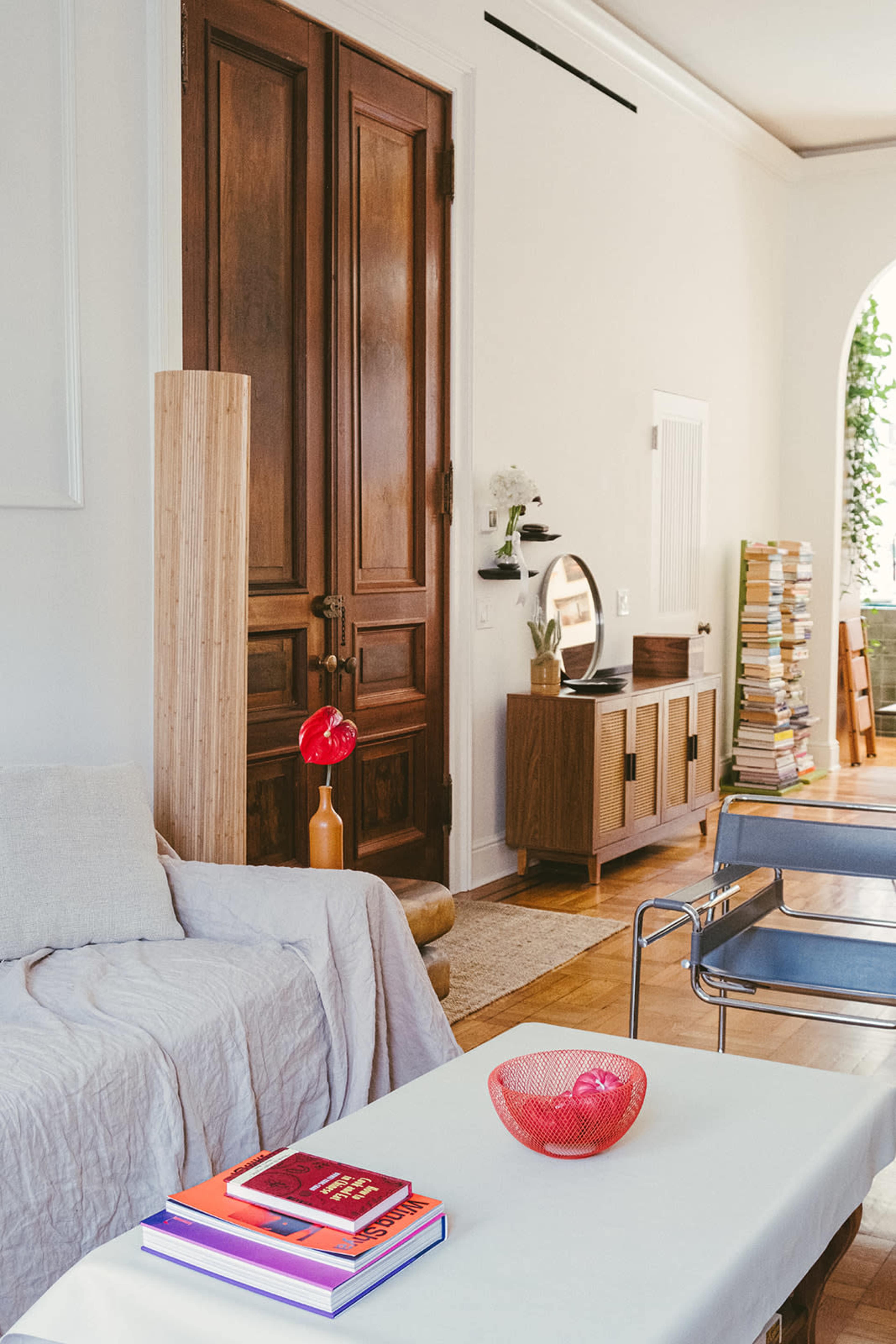 The image shows a modern living room with a couch, a coffee table topped with books and a decorative bowl, and wooden doors leading to another area, alongside a stack of magazines in the background.