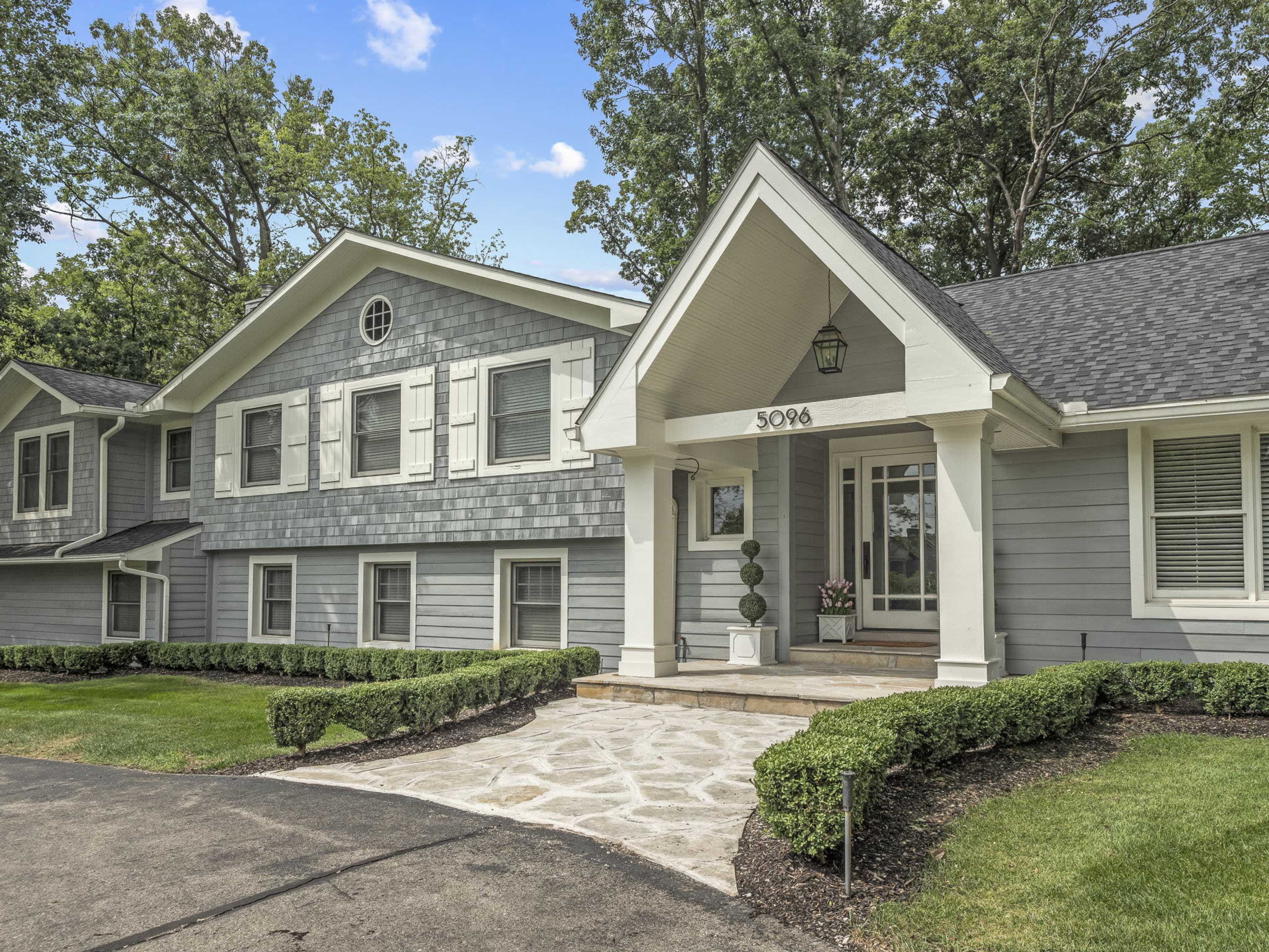 The image shows a gray, two-story house with a stone walkway leading to a covered front entrance, surrounded by neatly trimmed bushes and trees.