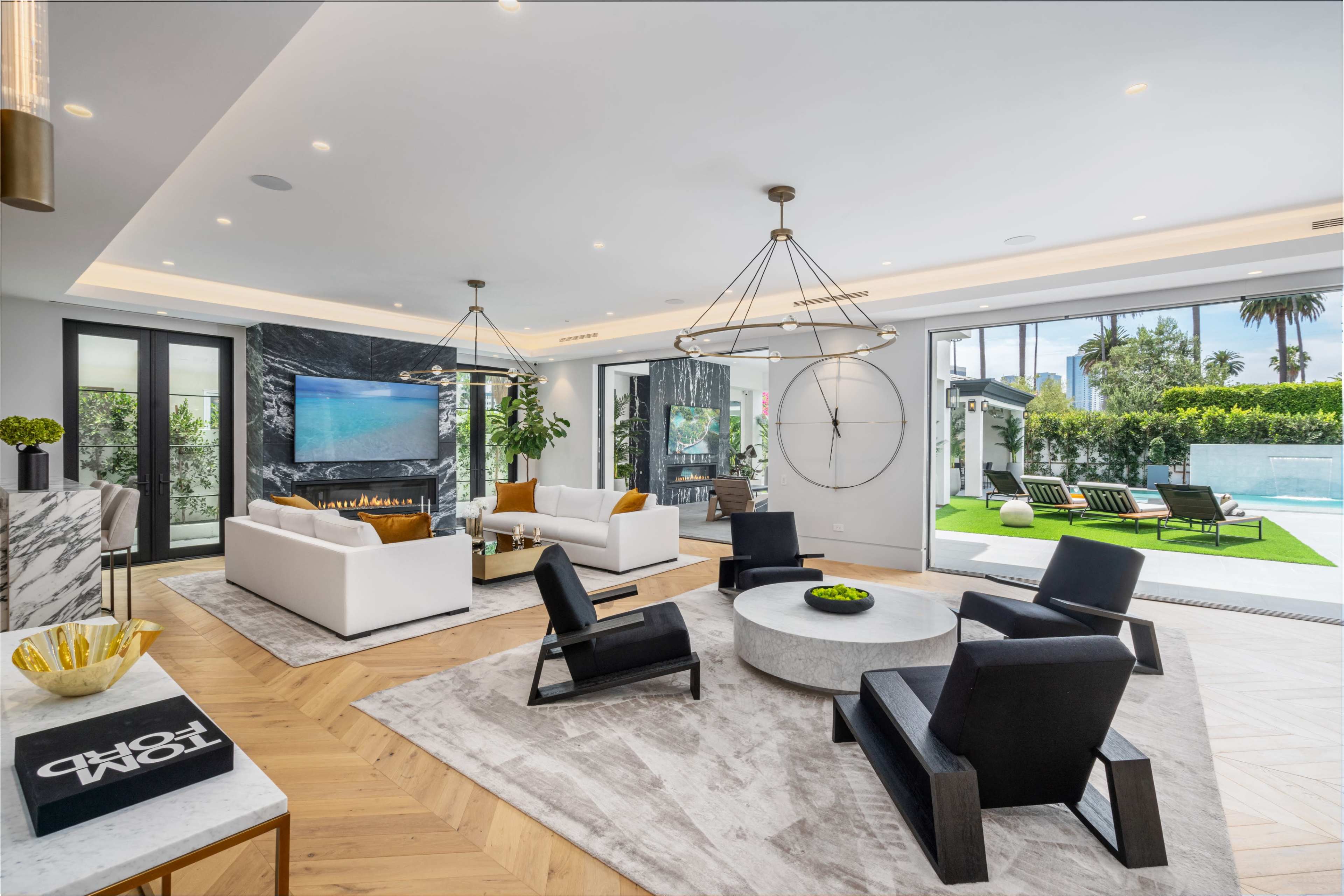 The image shows a modern living room with two white sofas, a circular coffee table, and black accent chairs, overlooking a landscaped outdoor area with a pool.