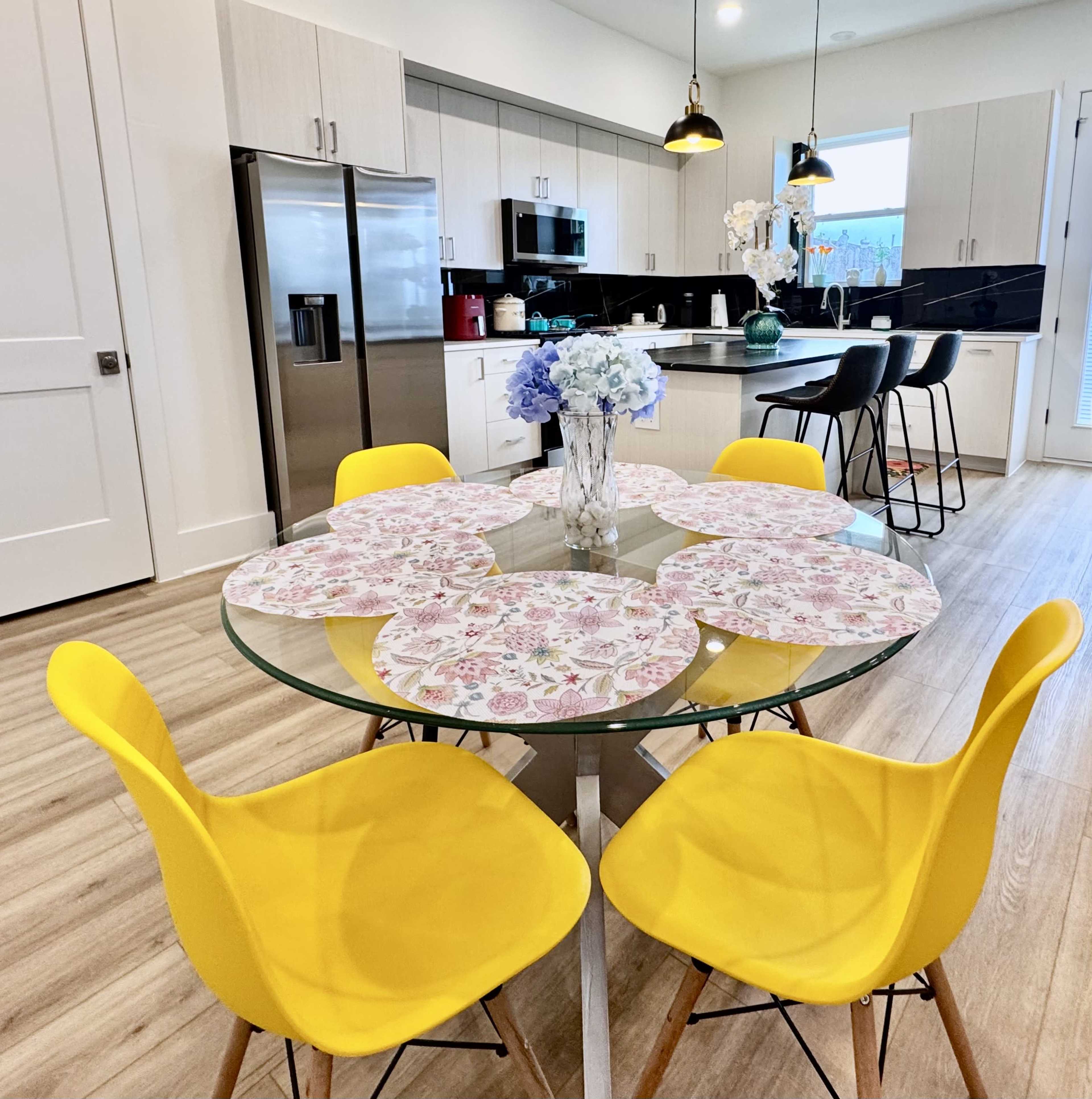 A round glass dining table with floral placemats is surrounded by yellow chairs in a modern kitchen featuring stainless steel appliances and white cabinetry.