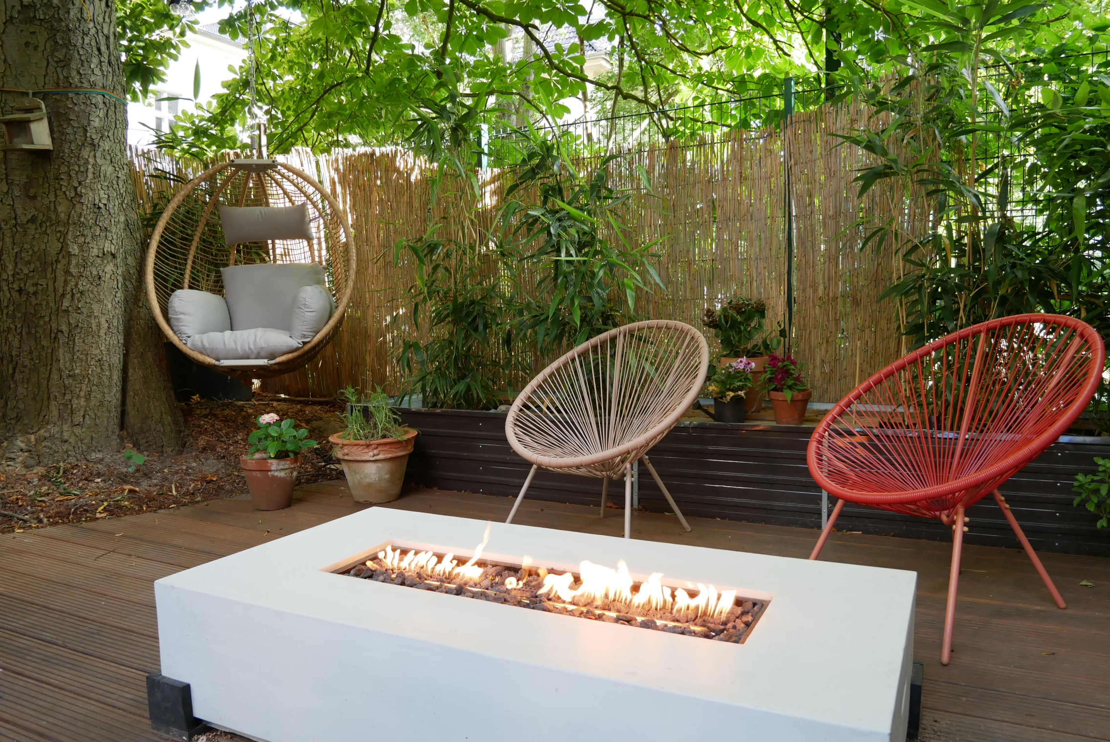 A modern outdoor seating area features a fire pit surrounded by two stylish chairs and a hanging swing, all amidst lush greenery.