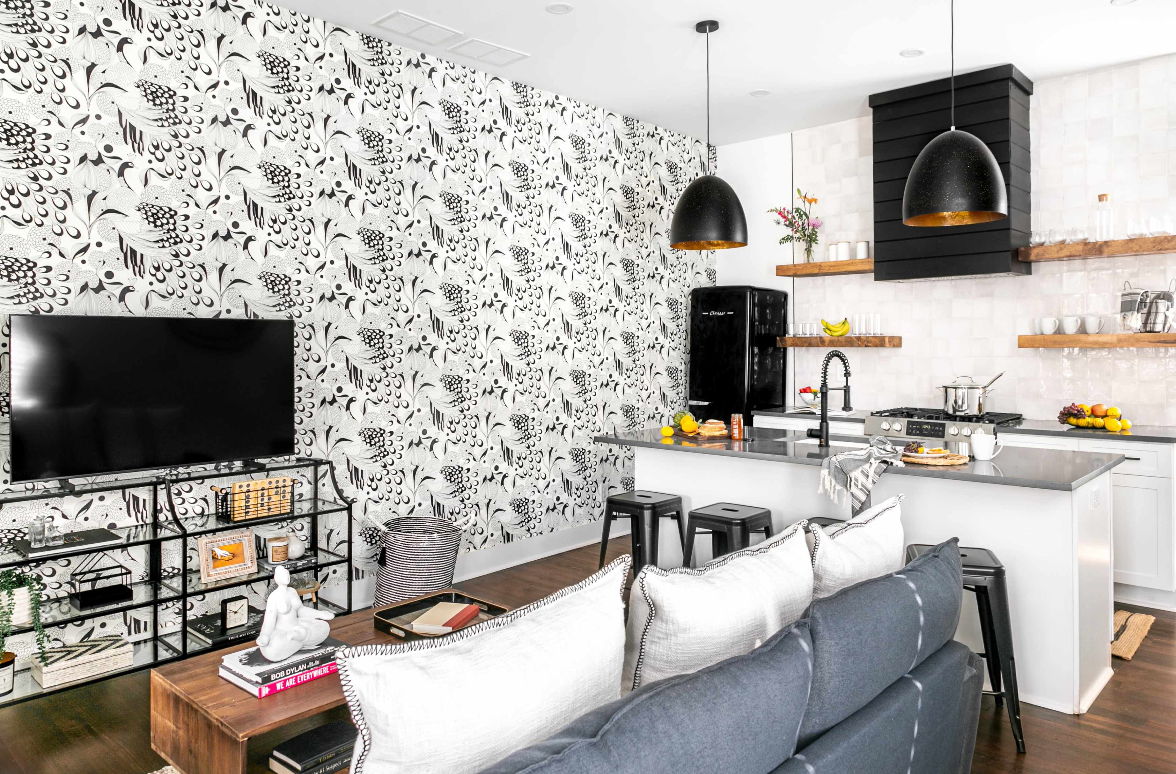 A modern living space featuring a black and white floral accent wall, a kitchen area with a bar counter, a dark sofa, and a television mounted on the wall.
