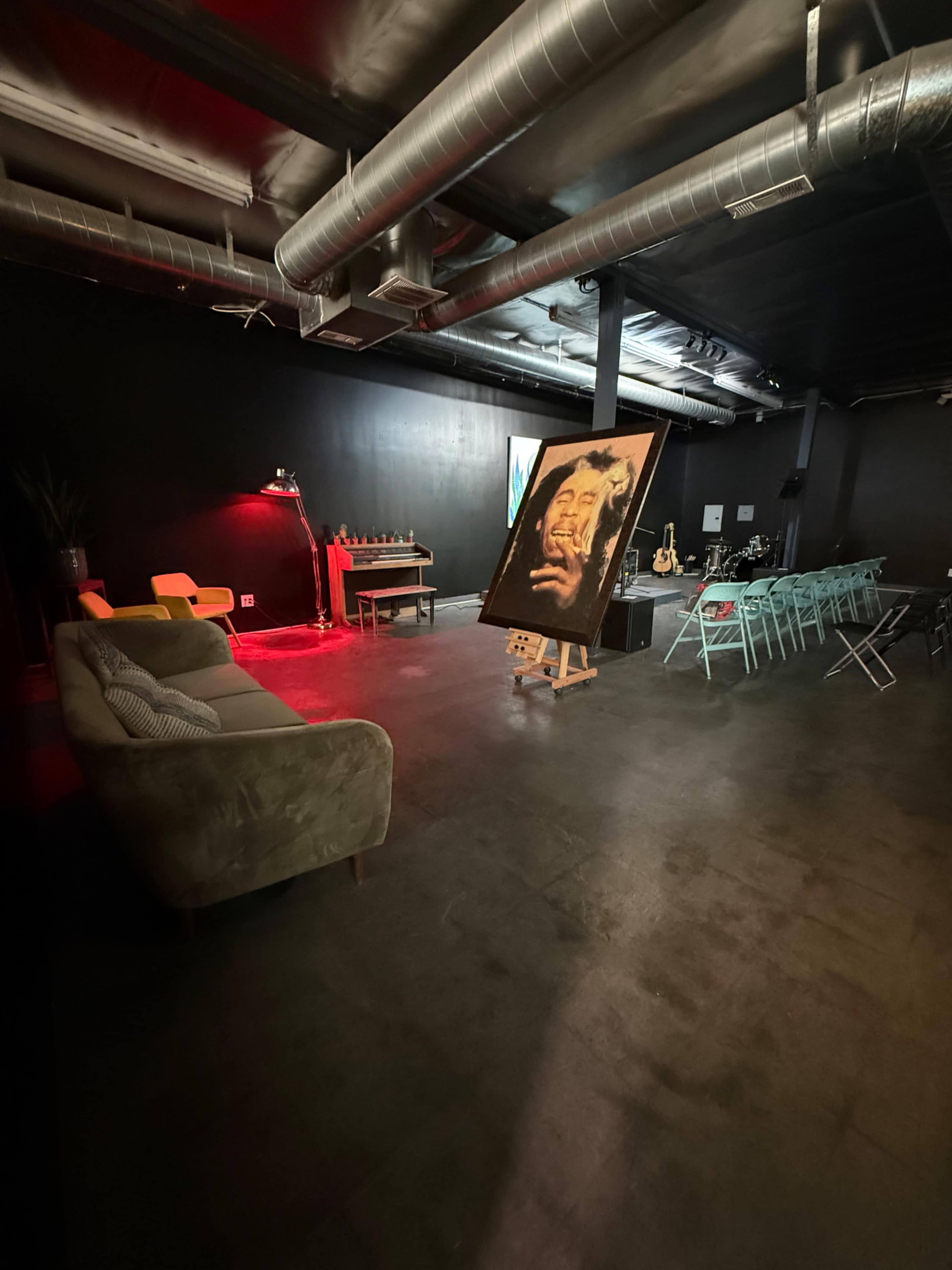 The image shows an indoor space with a large painting on an easel, a green couch, yellow chairs, and tables arranged for a gathering against a black wall.
