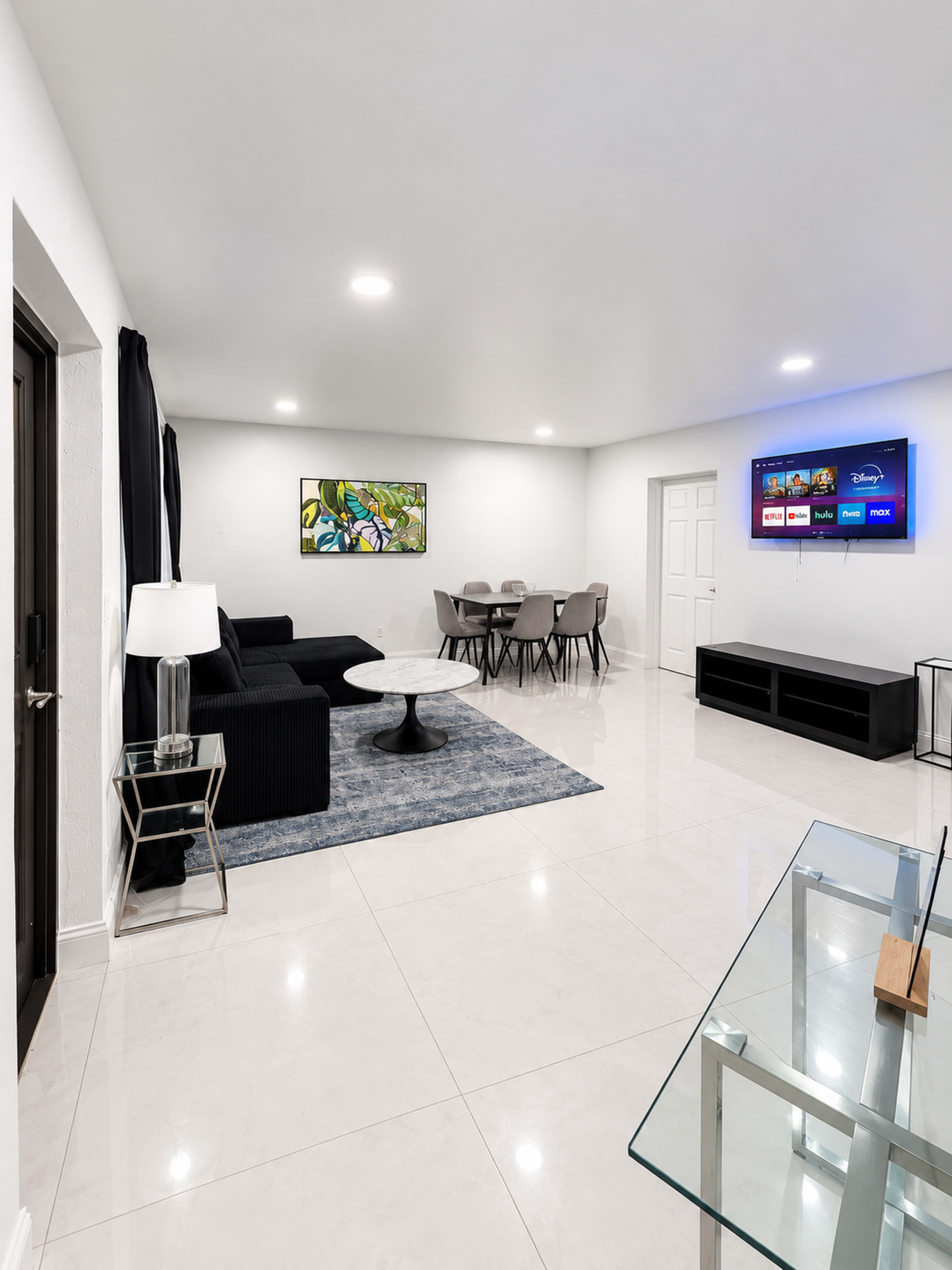 The image shows a modern living room with a black sofa, a round coffee table, a dining area with chairs, and a wall-mounted television.