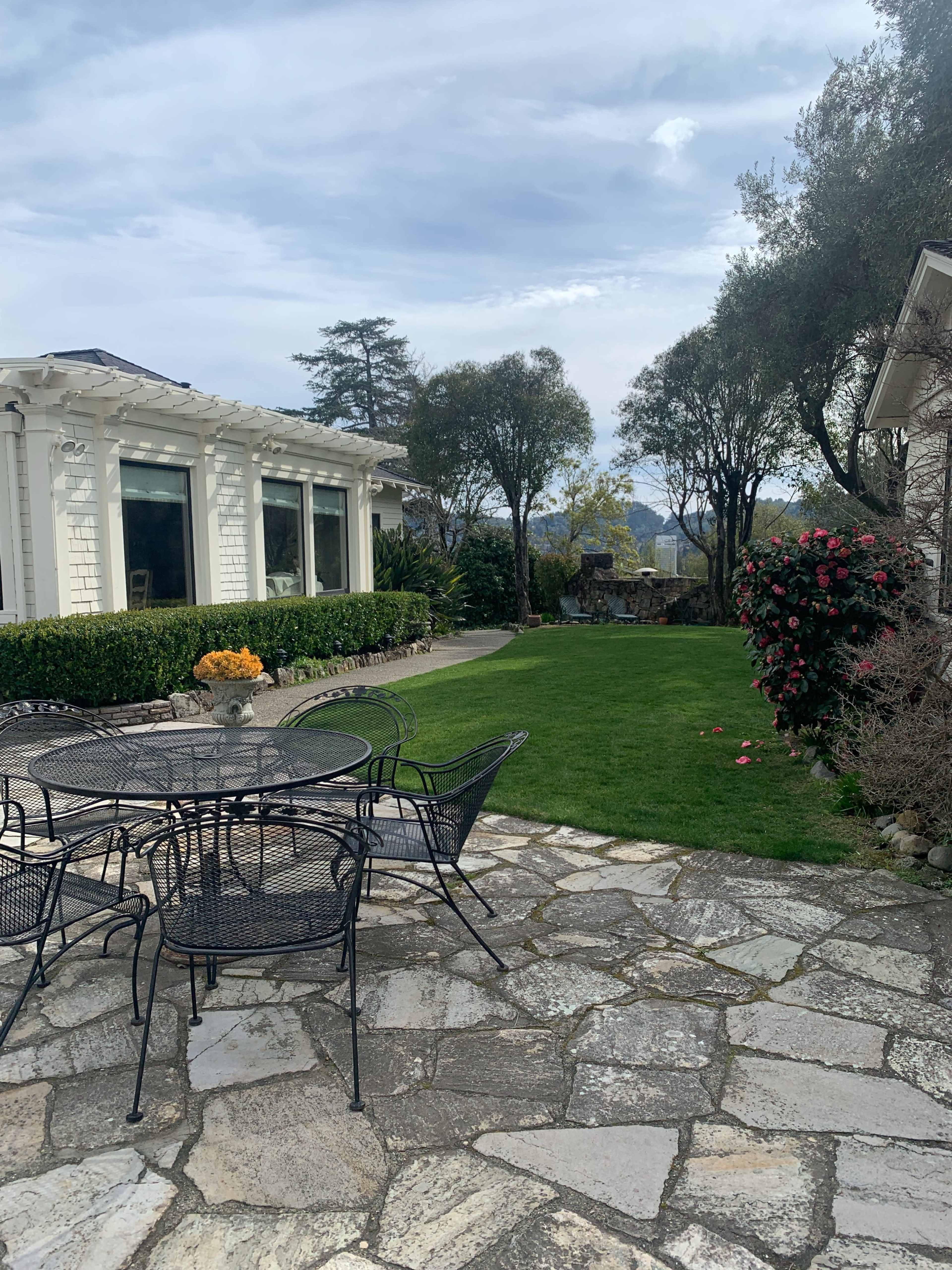 A stone patio with black metal furniture overlooks a well-maintained lawn and garden area beside a large white house.