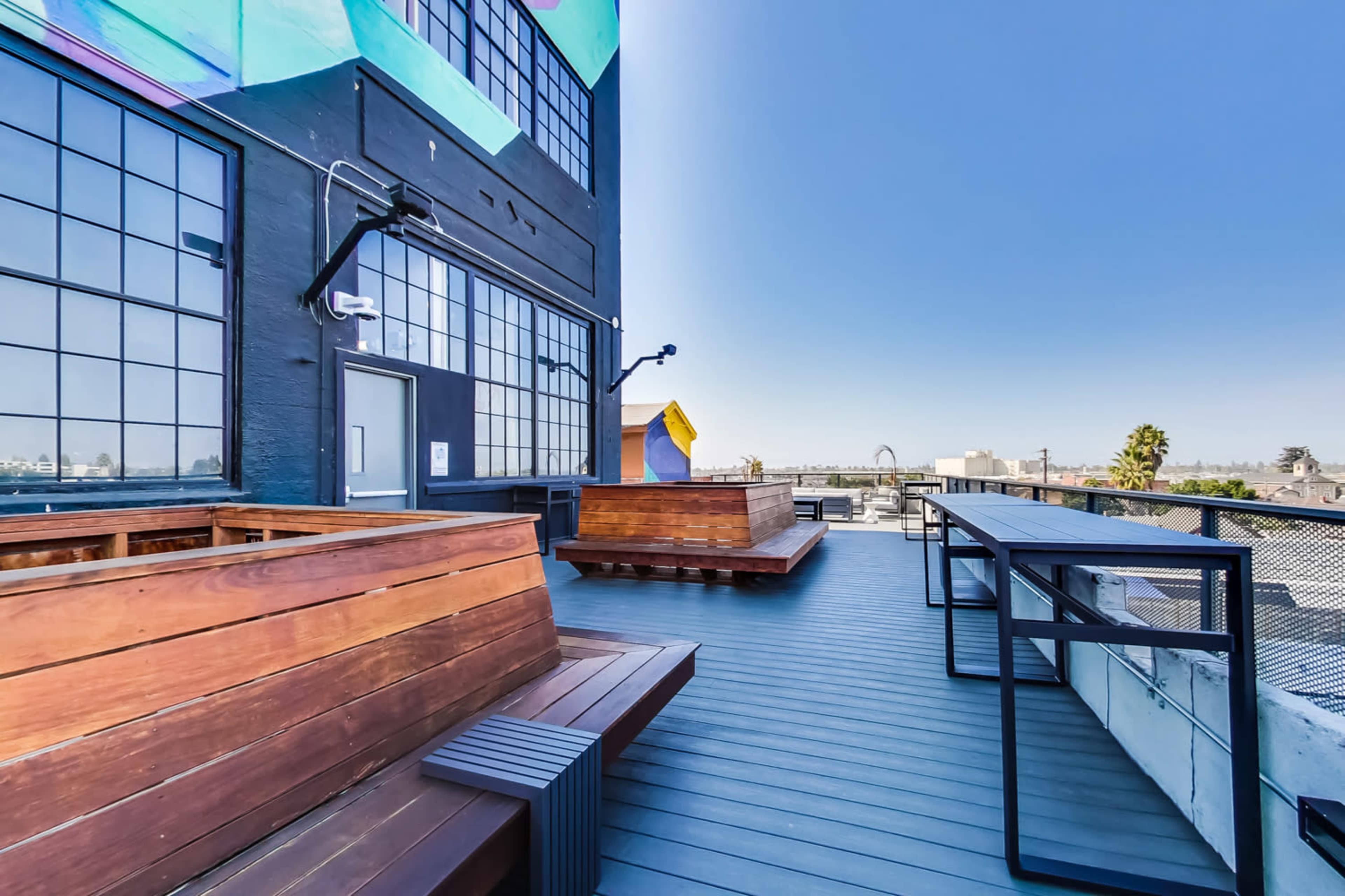 A rooftop area with wooden seating and tables, overlooking a clear sky and distant structures.