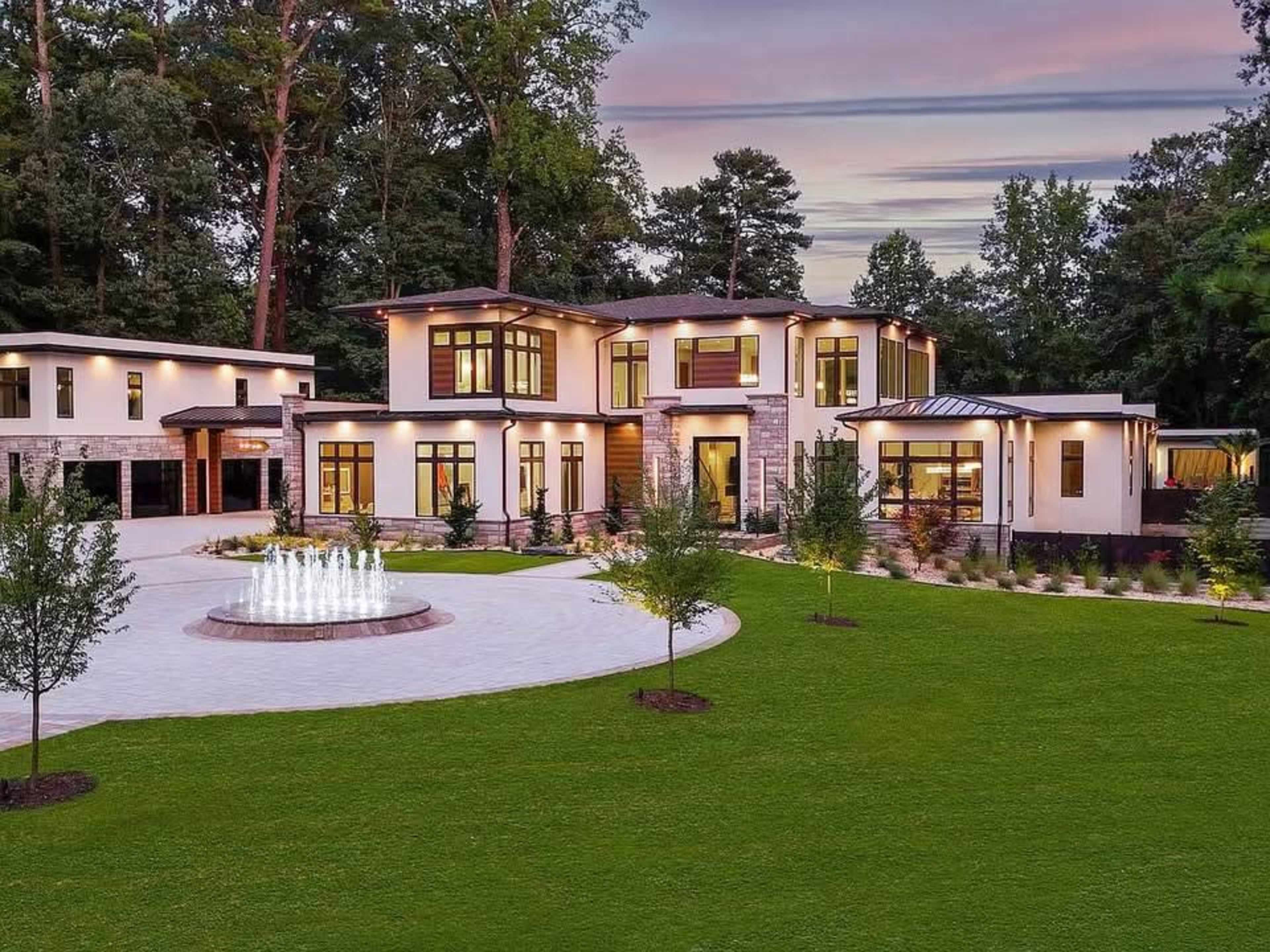 A modern two-story house with large windows and a fountain is surrounded by manicured lawns and trees.