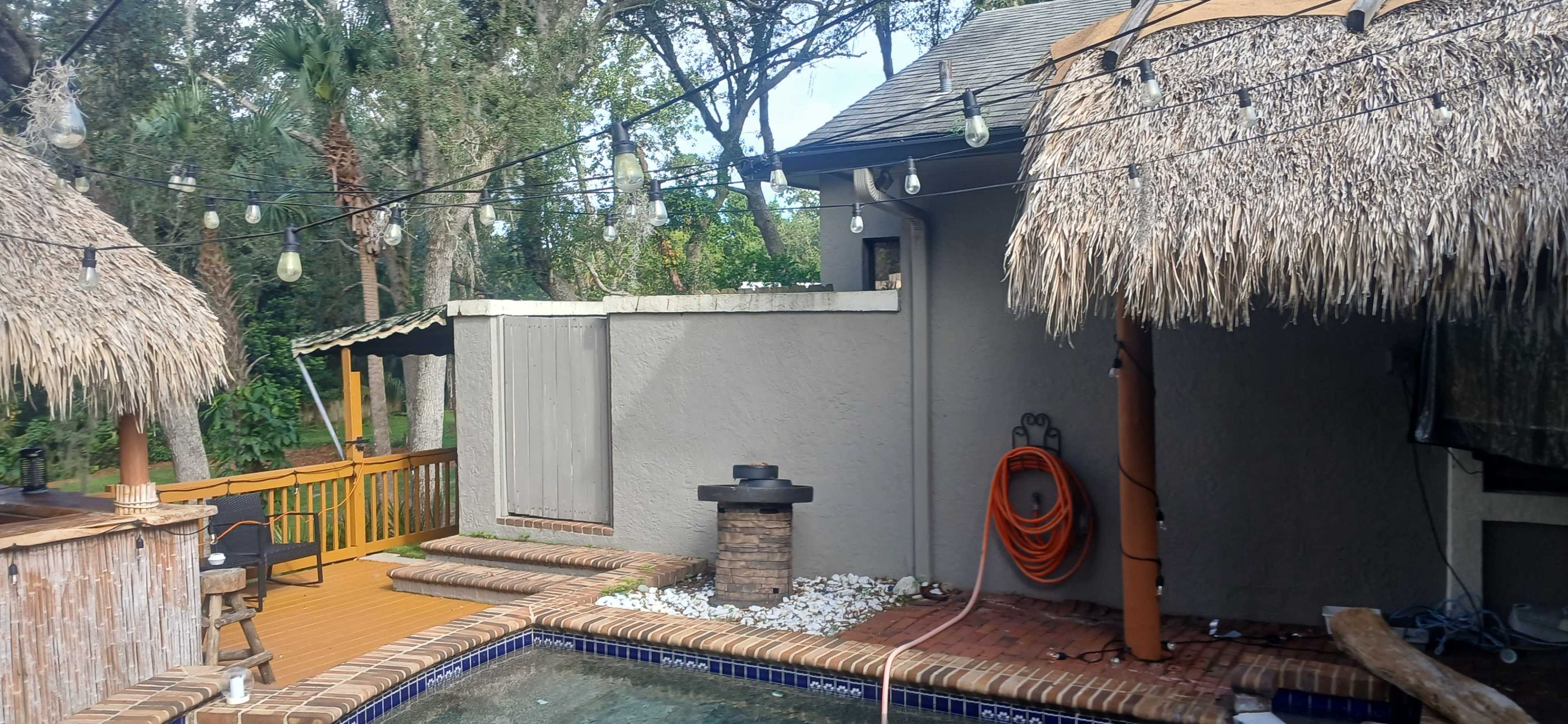The image shows a backyard scene with a swimming pool, wooden deck, and a thatched-roof structure, alongside a decorative stone feature and hanging lights.