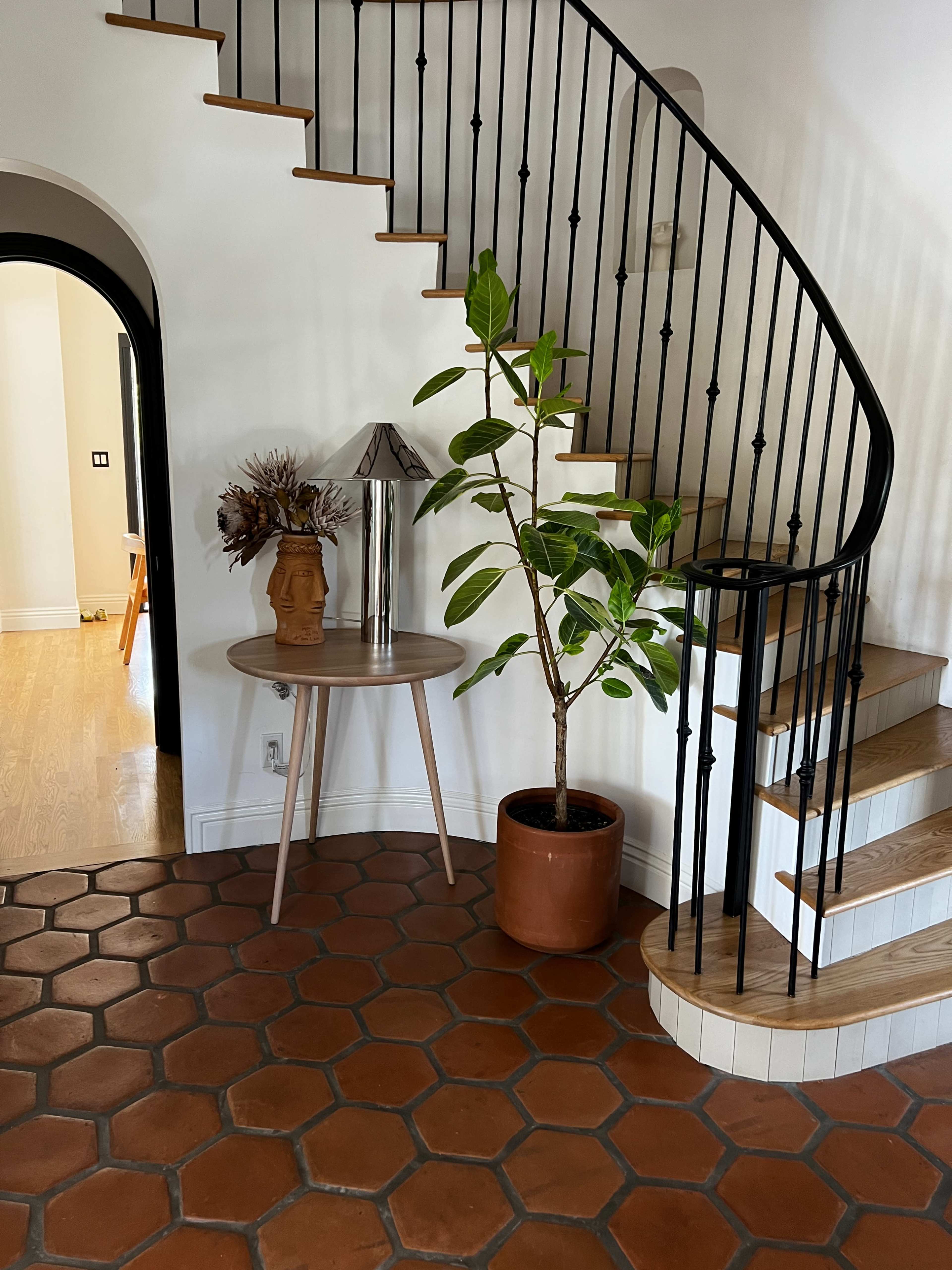A curved staircase with black railing rises next to a round table featuring a decorative lamp and a plant in an orange pot, set against a backdrop of terracotta tiled flooring.