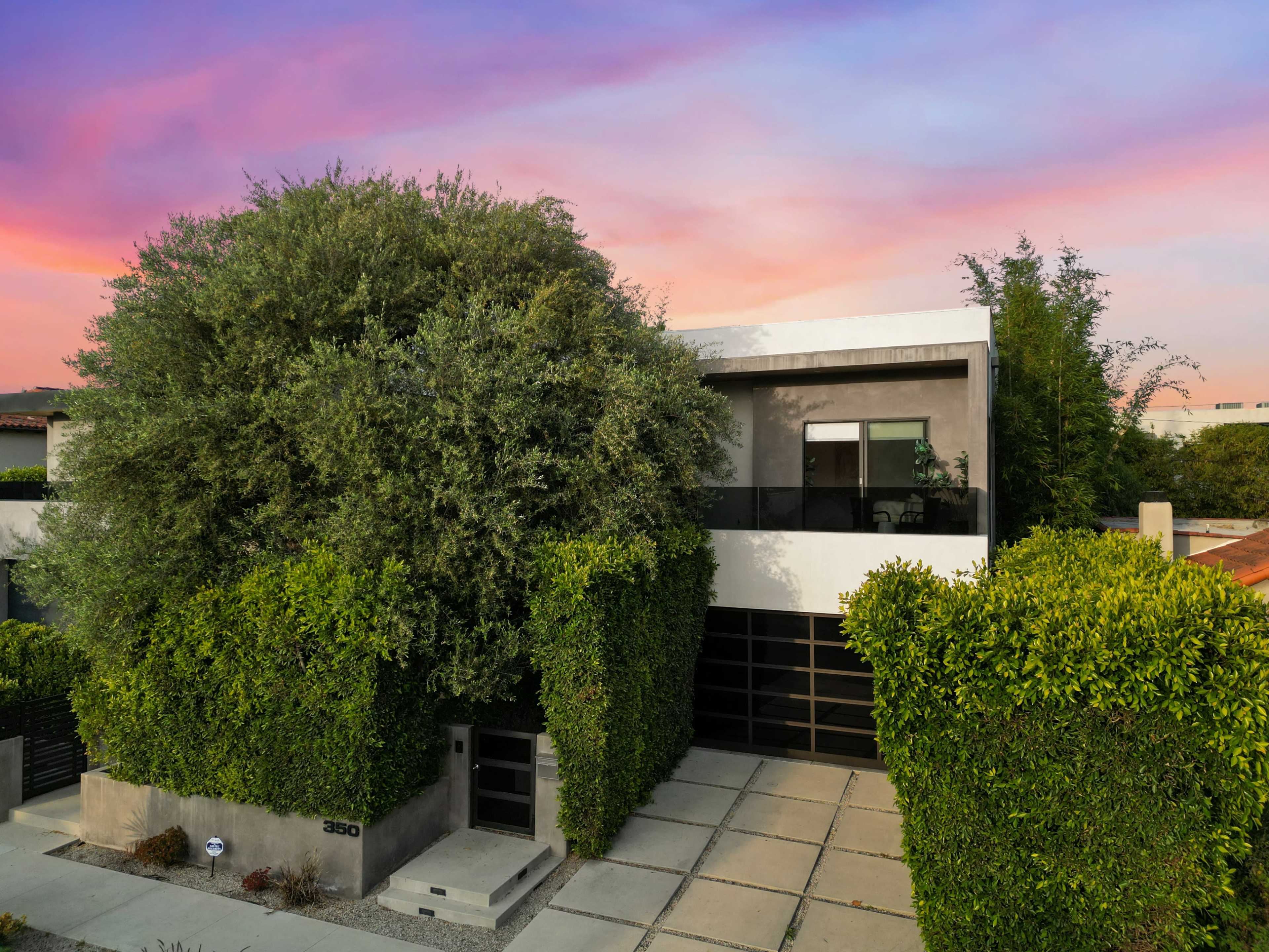A modern two-story house is set back behind tall greenery, with a driveway leading to a garage on the front.
