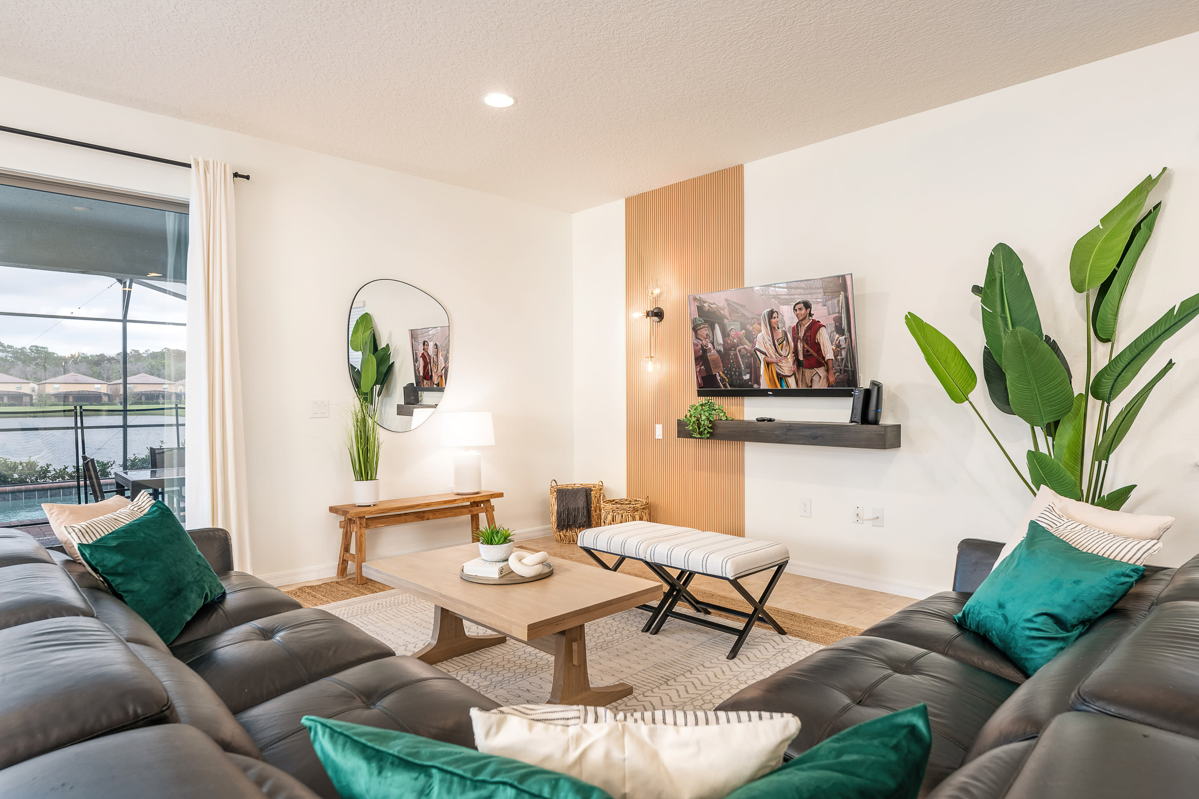 The living room features two dark leather sofas, a wooden coffee table, a large wall-mounted television, and decorative plants, all arranged around a light-colored area rug.