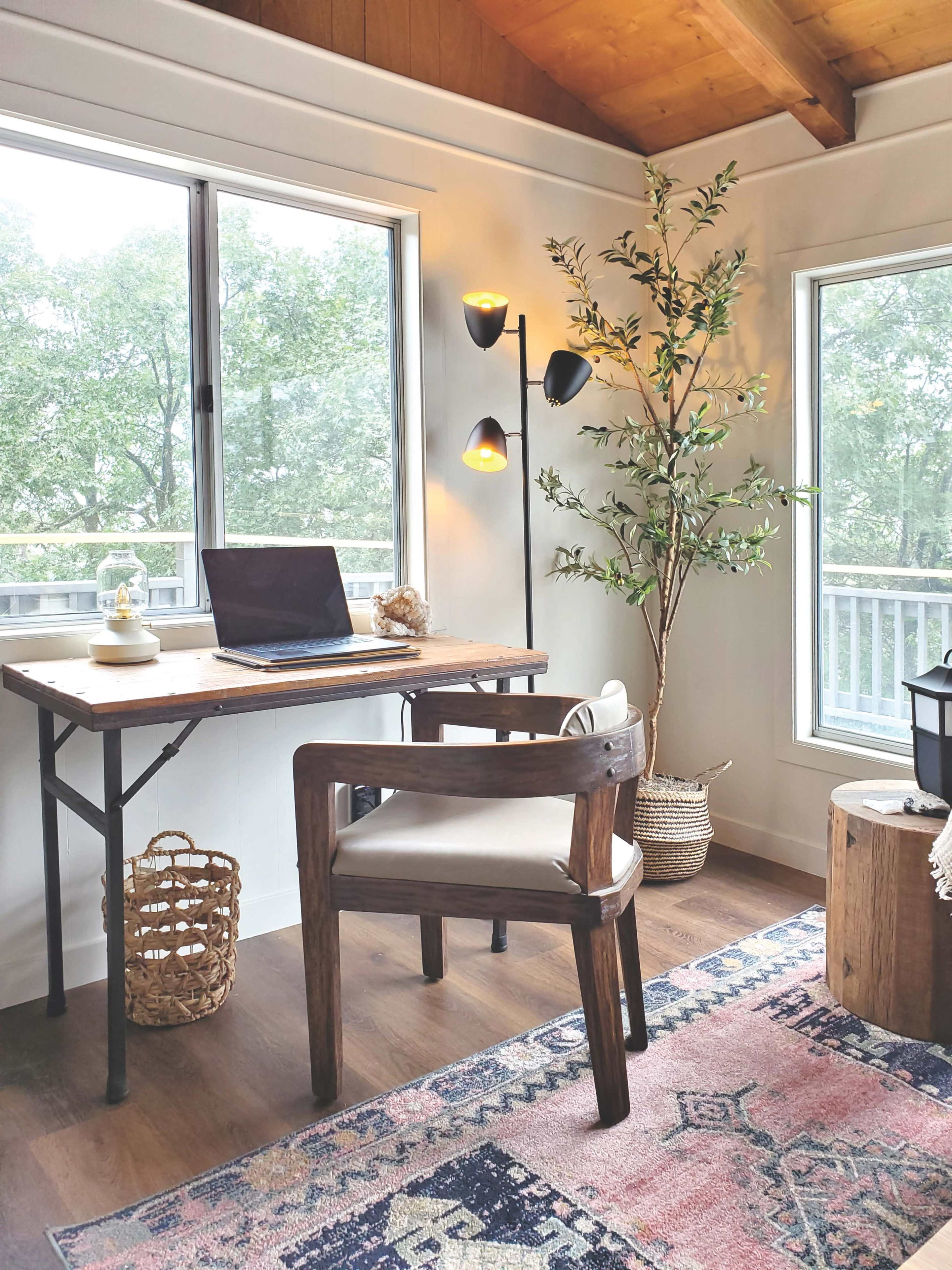A workspace with a wooden desk, a laptop, a chair, and a potted plant, located near large windows with natural light.