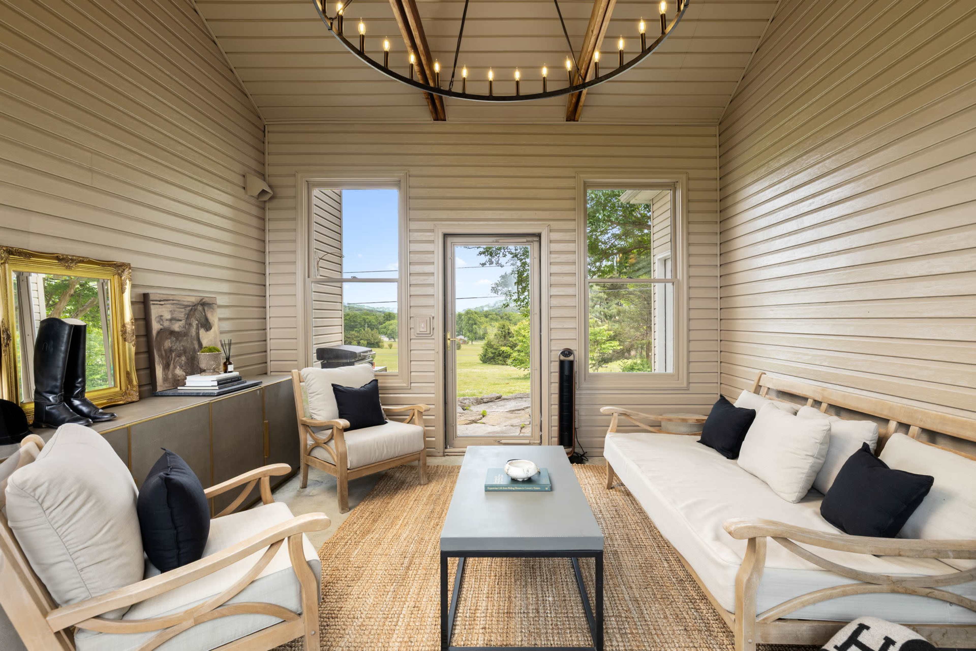 The image shows a light-filled sunroom with wooden walls, two sofas covered in white and black pillows, a coffee table in the center, and large windows offering a view of the outdoors.