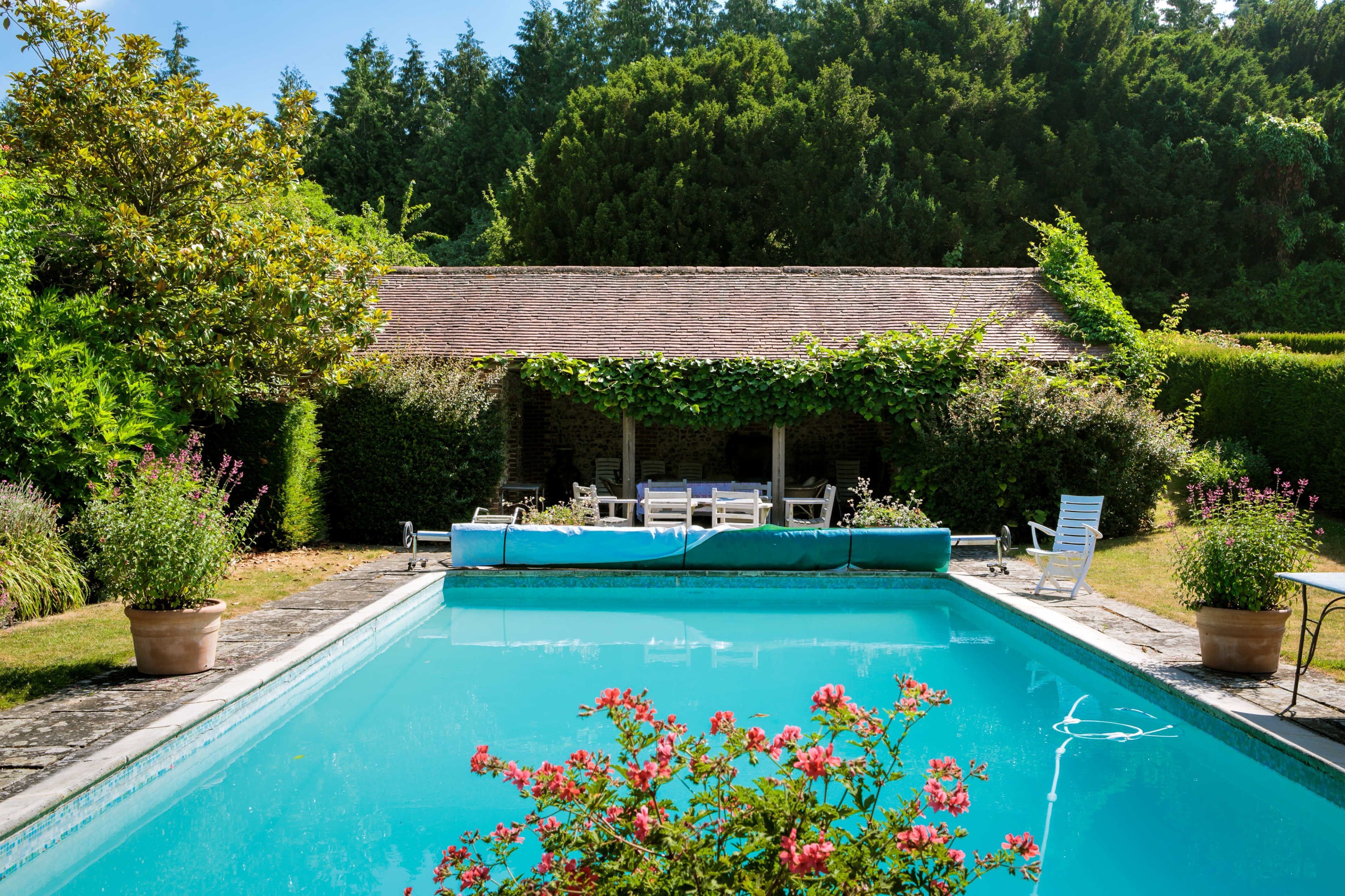 A swimming pool with blue water is surrounded by landscaping, featuring a covered seating area and potted plants.