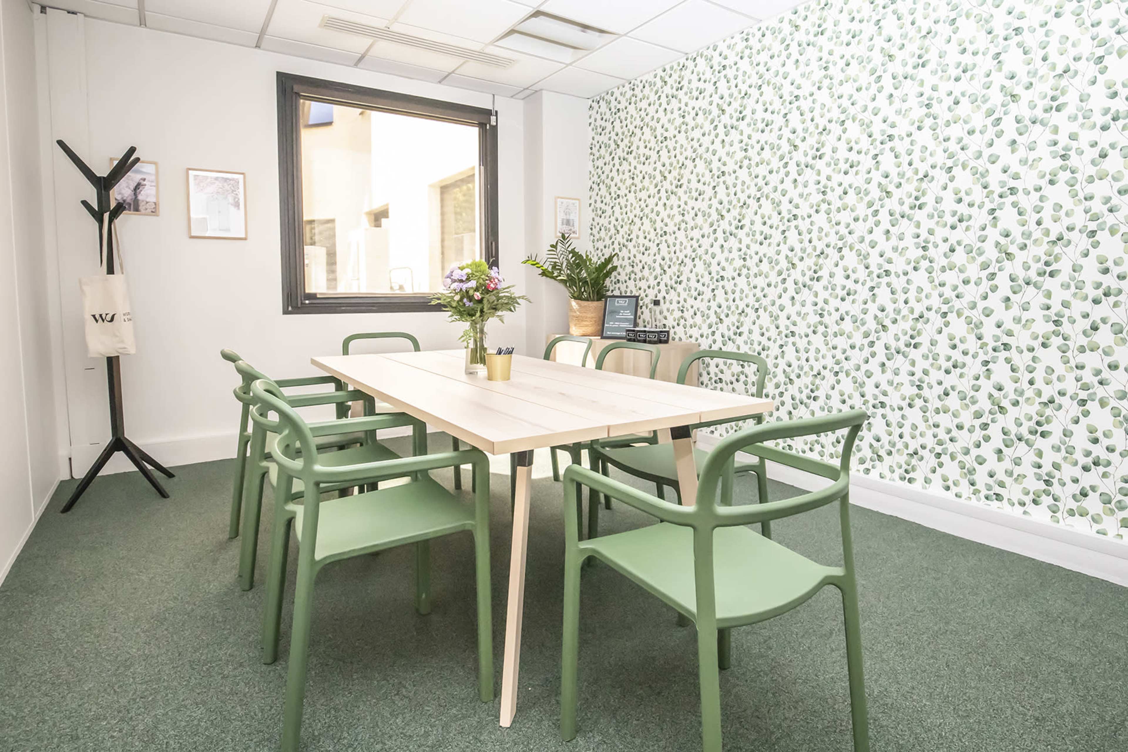 A bright meeting room with a large wooden table surrounded by green chairs, and a wall decorated with a leafy pattern.