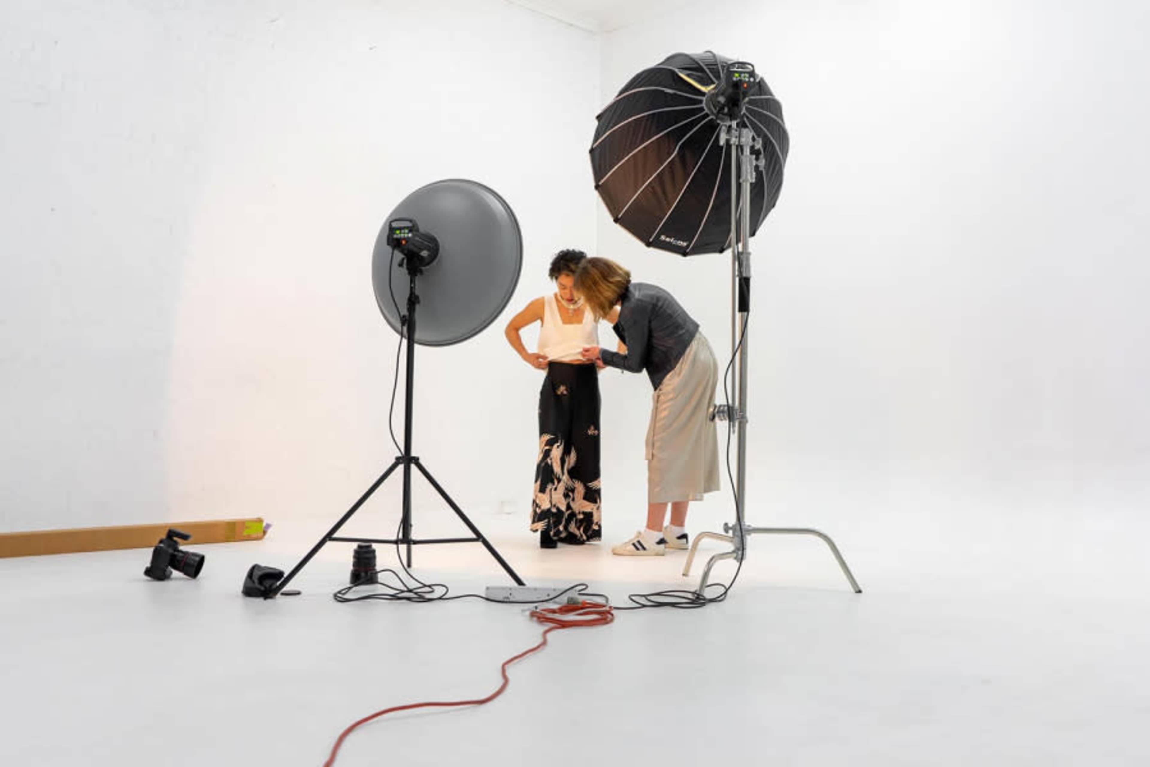 Two women are adjusting clothing in a well-lit photography studio featuring large softbox lights and a seamless white backdrop.