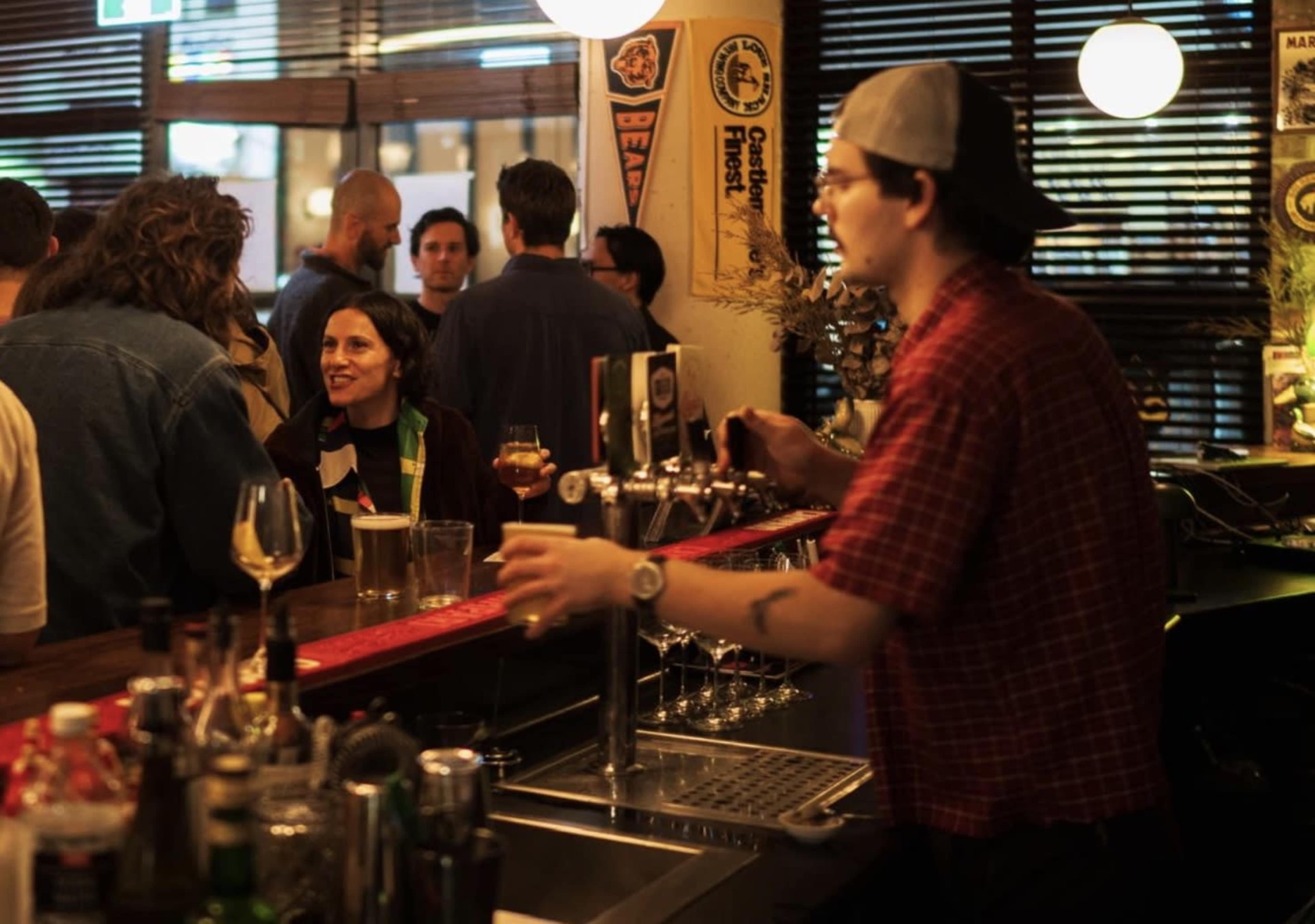 A bartender in a checked shirt pours a drink at a bustling bar filled with patrons engaged in conversation.