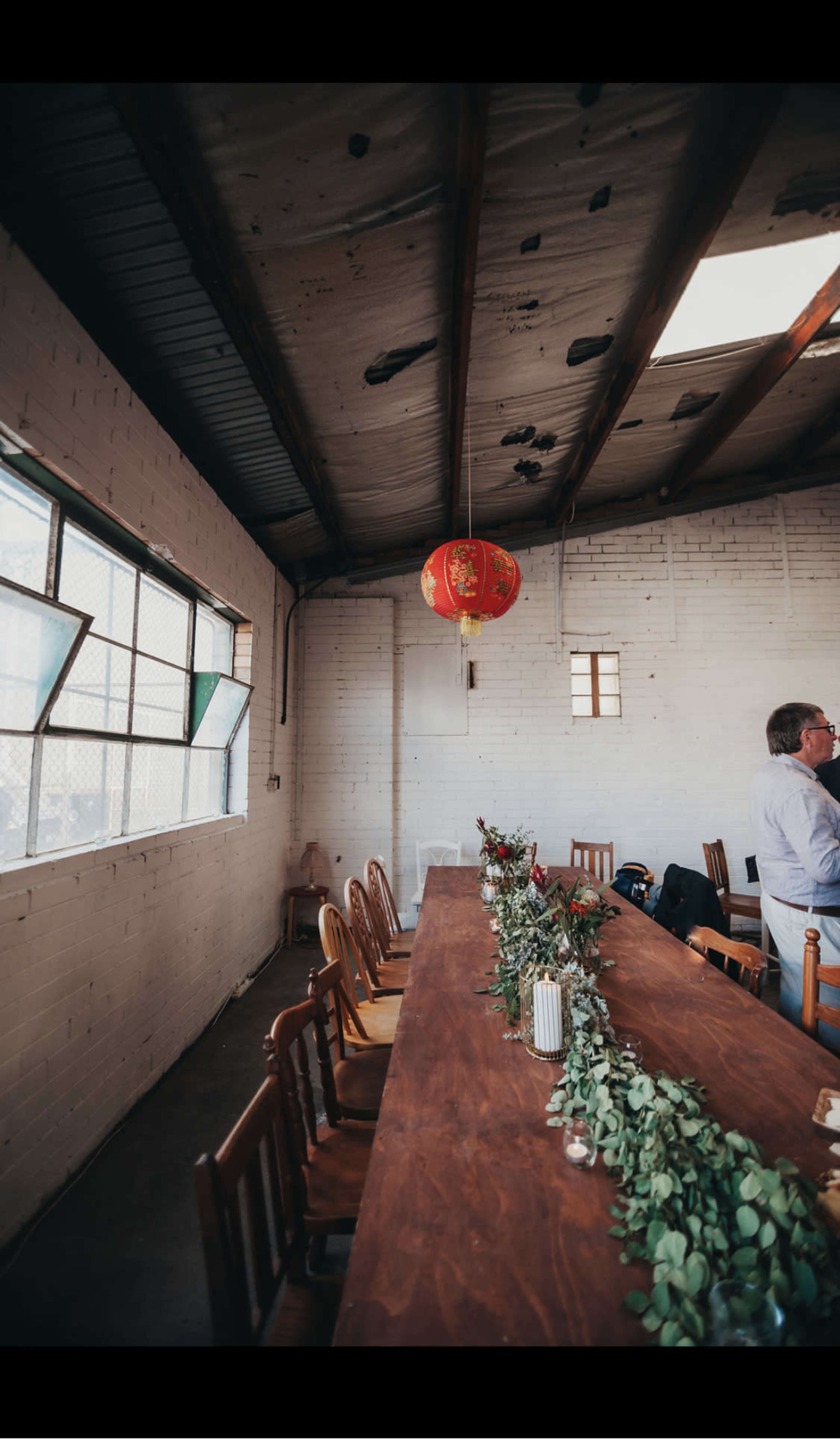 The image shows a rustic room with a long wooden table adorned with greenery and flowers, under a red lantern hanging from the ceiling.