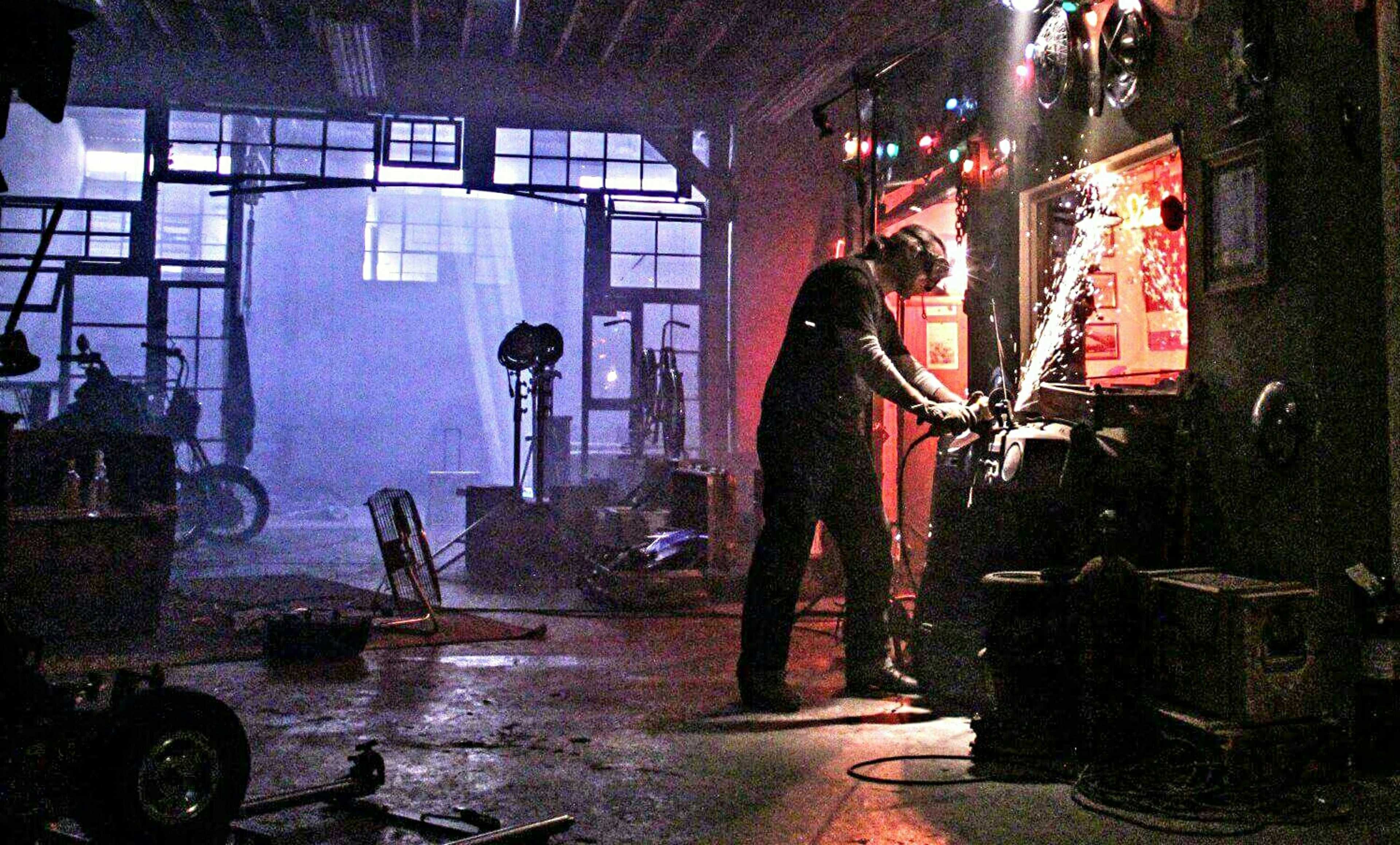 A man works on a metal project in a dimly lit workshop, surrounded by various tools and equipment.