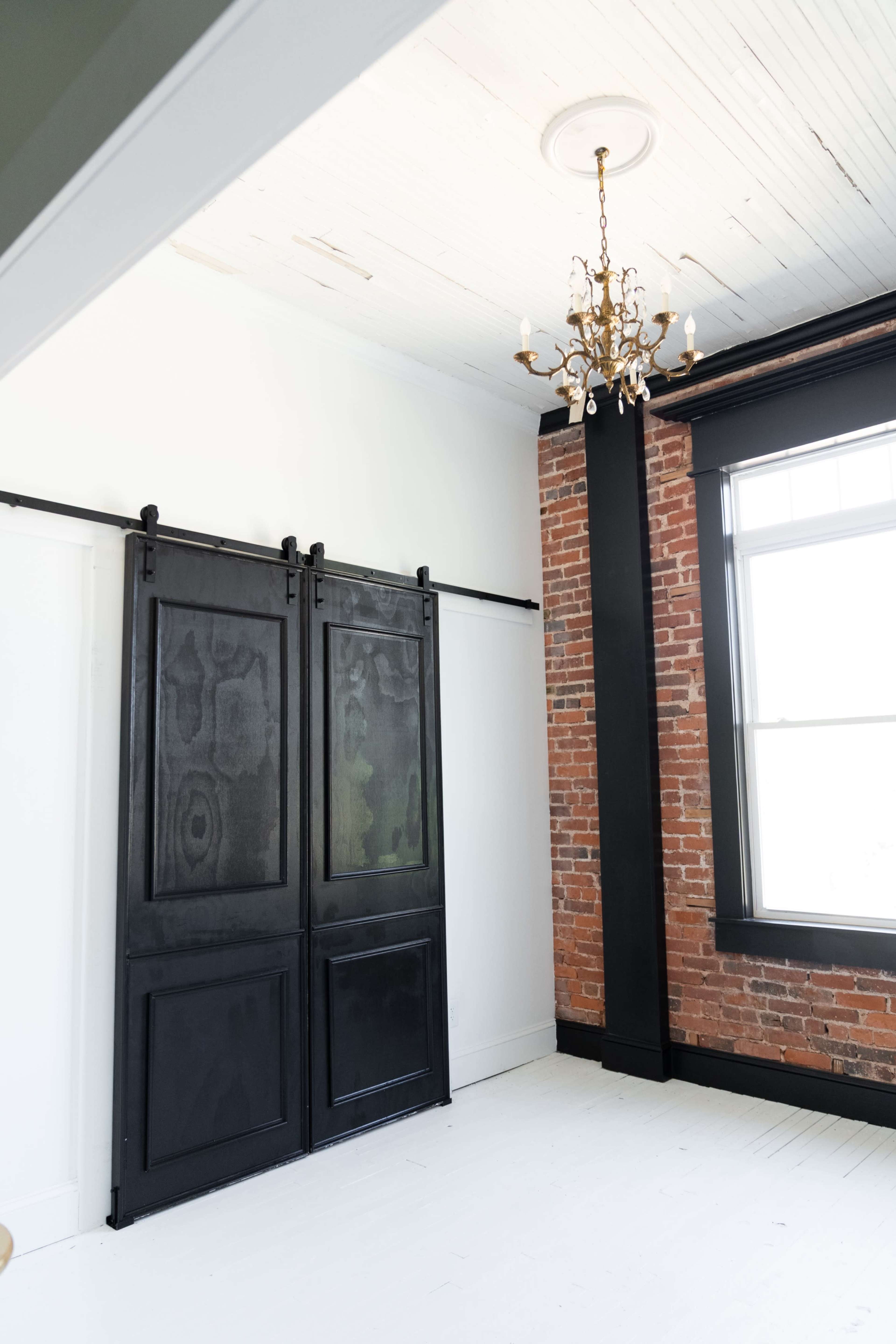 A black sliding barn door is installed on a wall next to a brick column in a brightly lit room with a chandelier.