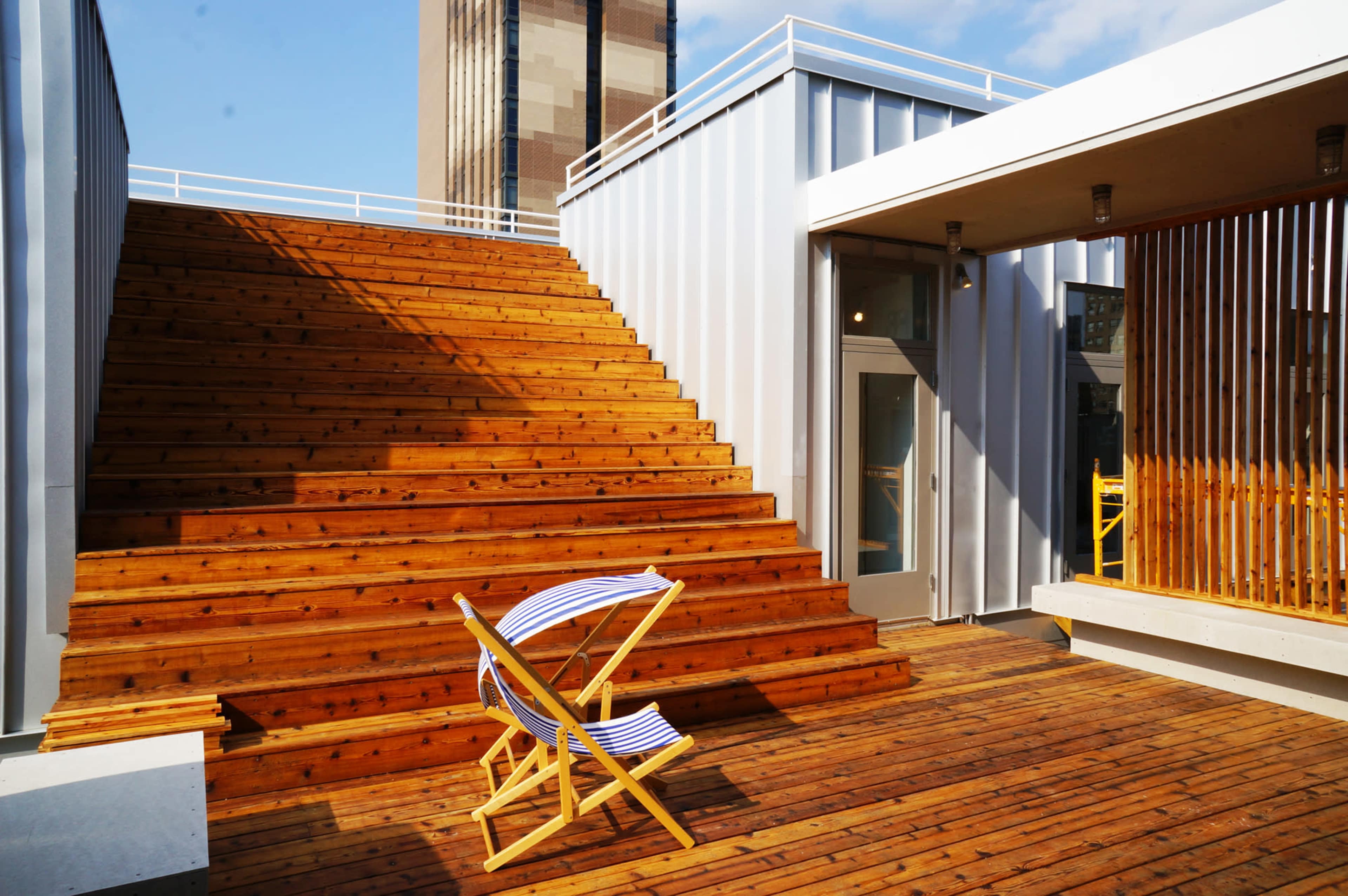 A wooden staircase leading up to a rooftop area, flanked by modern metal structures and featuring a single deck chair on the wooden floor.