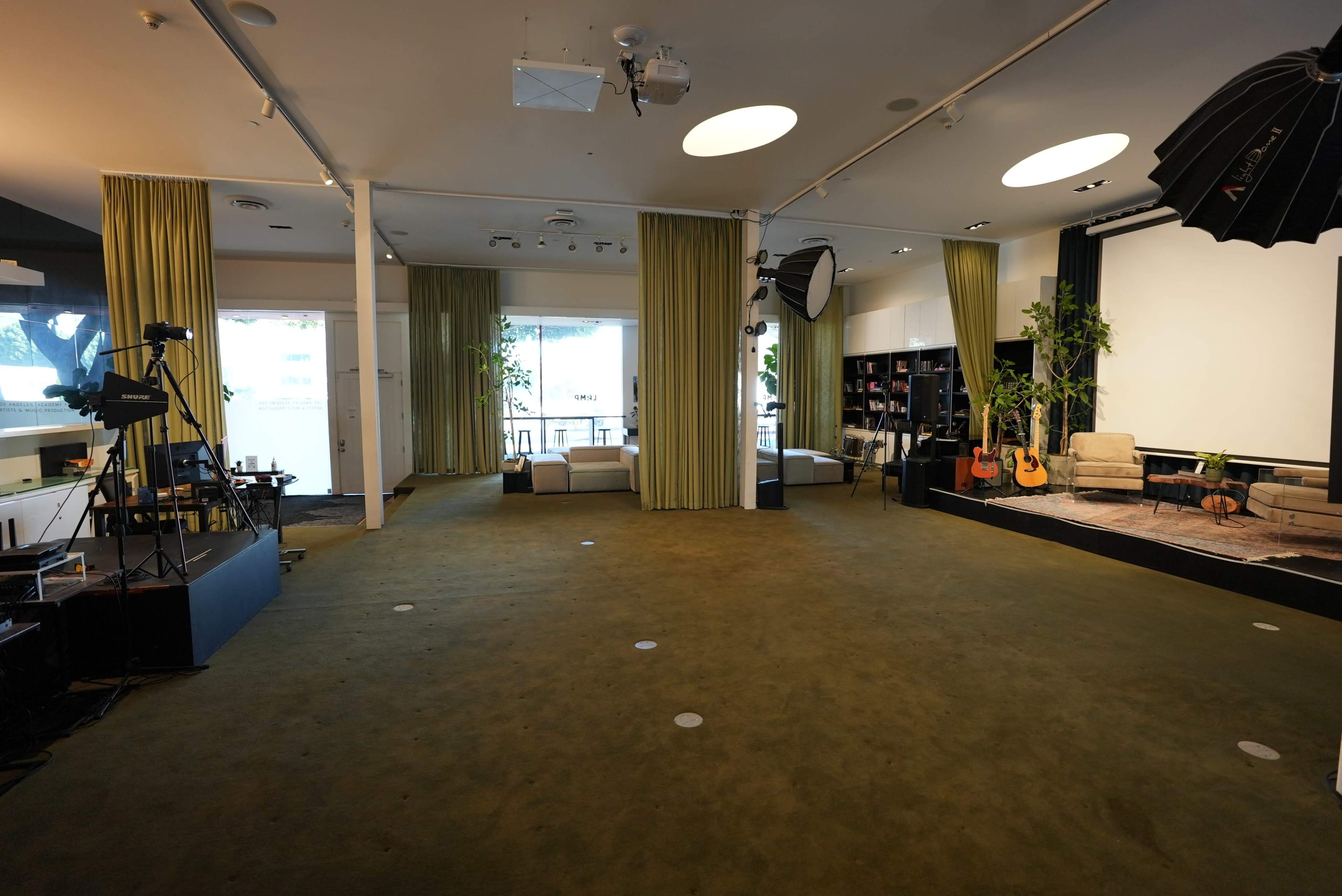 The image shows a spacious, empty studio with green carpet, large windows, and a seating area featuring couches, plants, and musical instruments.