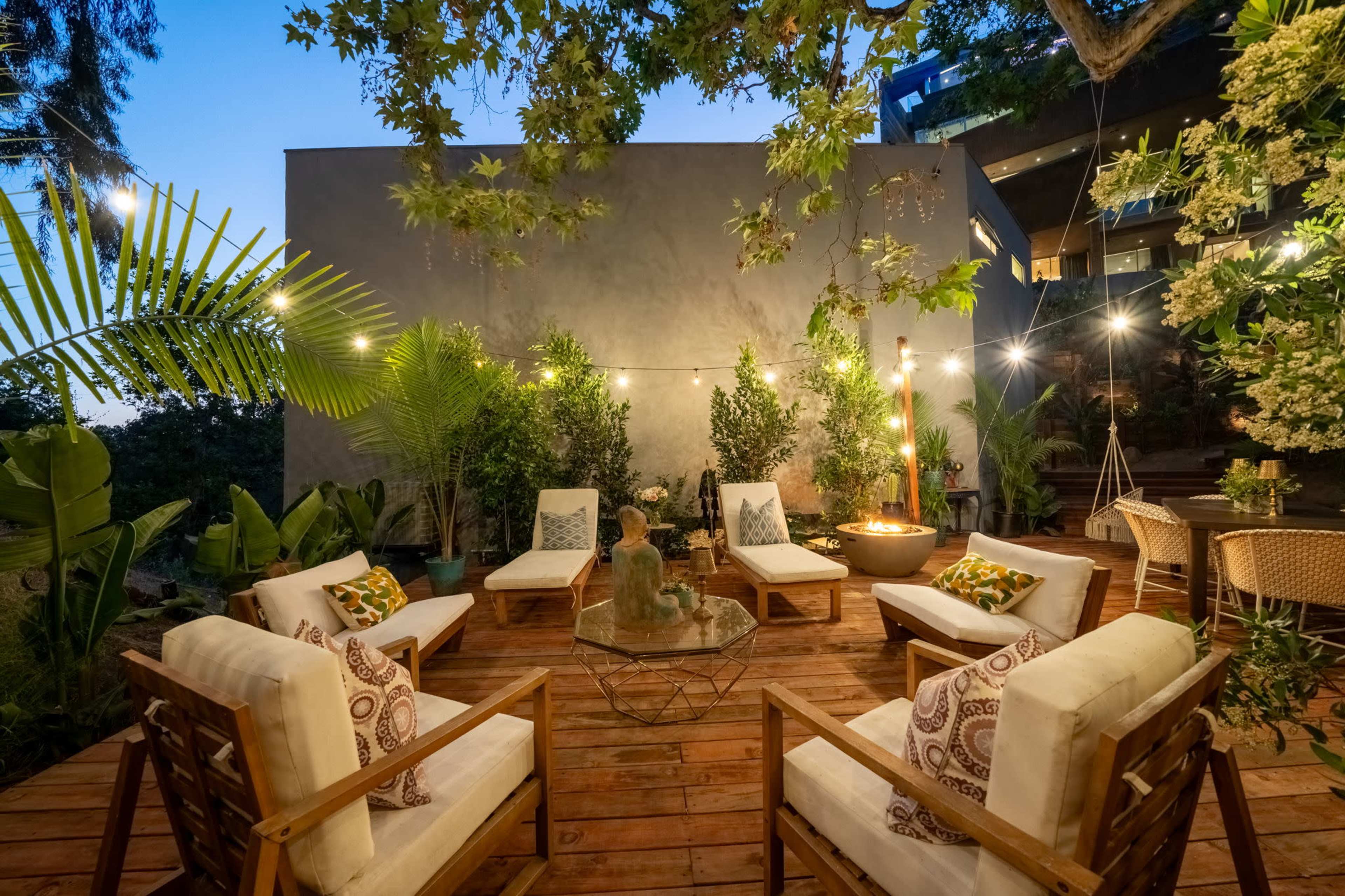 A cozy outdoor seating area with white chairs, a coffee table, and tropical plants, illuminated by string lights.