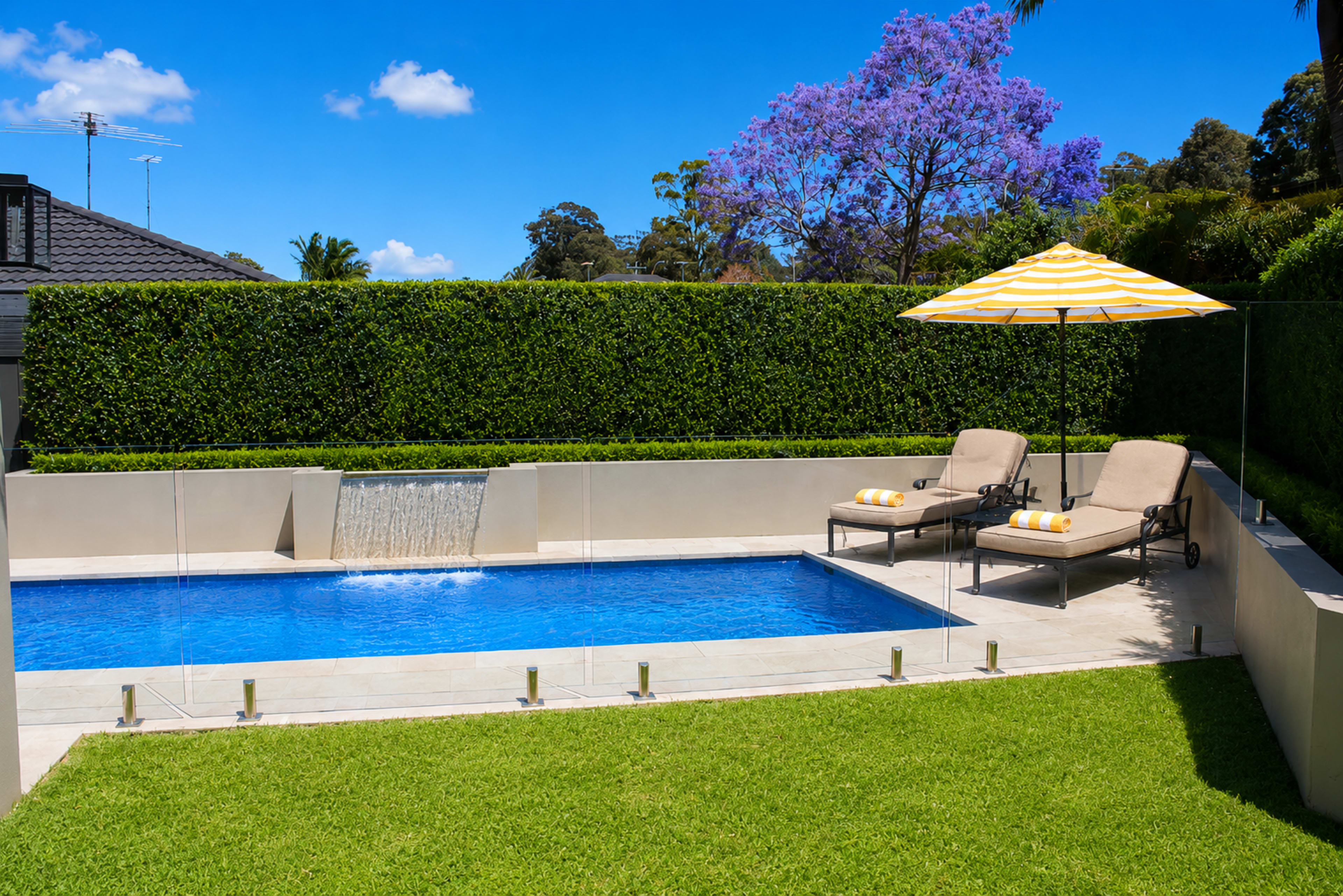 The image features a rectangular swimming pool surrounded by a grass lawn, with two loungers and a yellow-striped umbrella beside it, and a cascading water feature at one end.