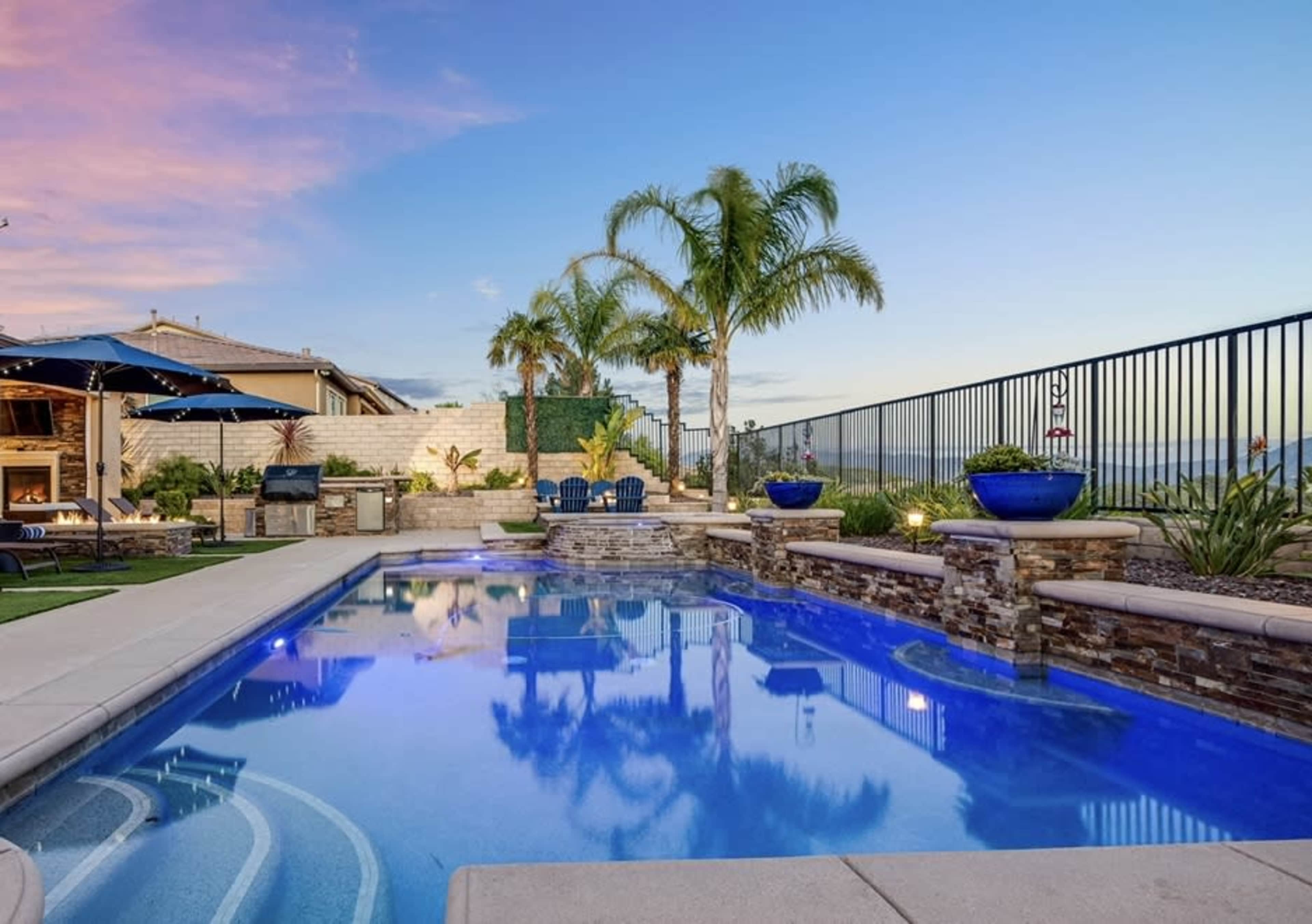 A backyard swimming pool is surrounded by palm trees, stone walls, and lounge chairs, with patio umbrellas and planters visible in the landscaped area.