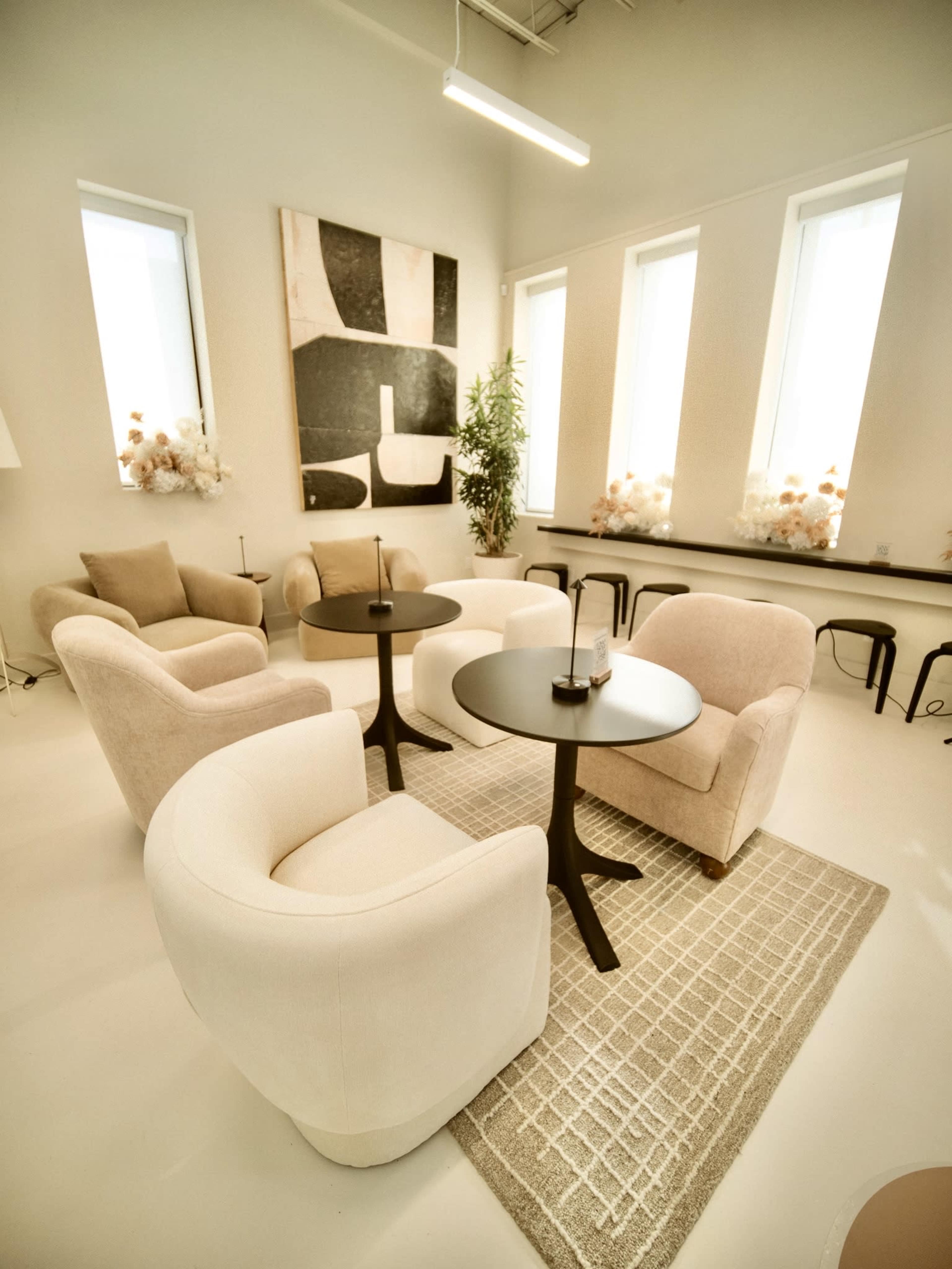 The image shows a modern, minimalist lounge area with two light-colored armchairs and a small round table positioned on a textured rug, illuminated by natural light from large windows.