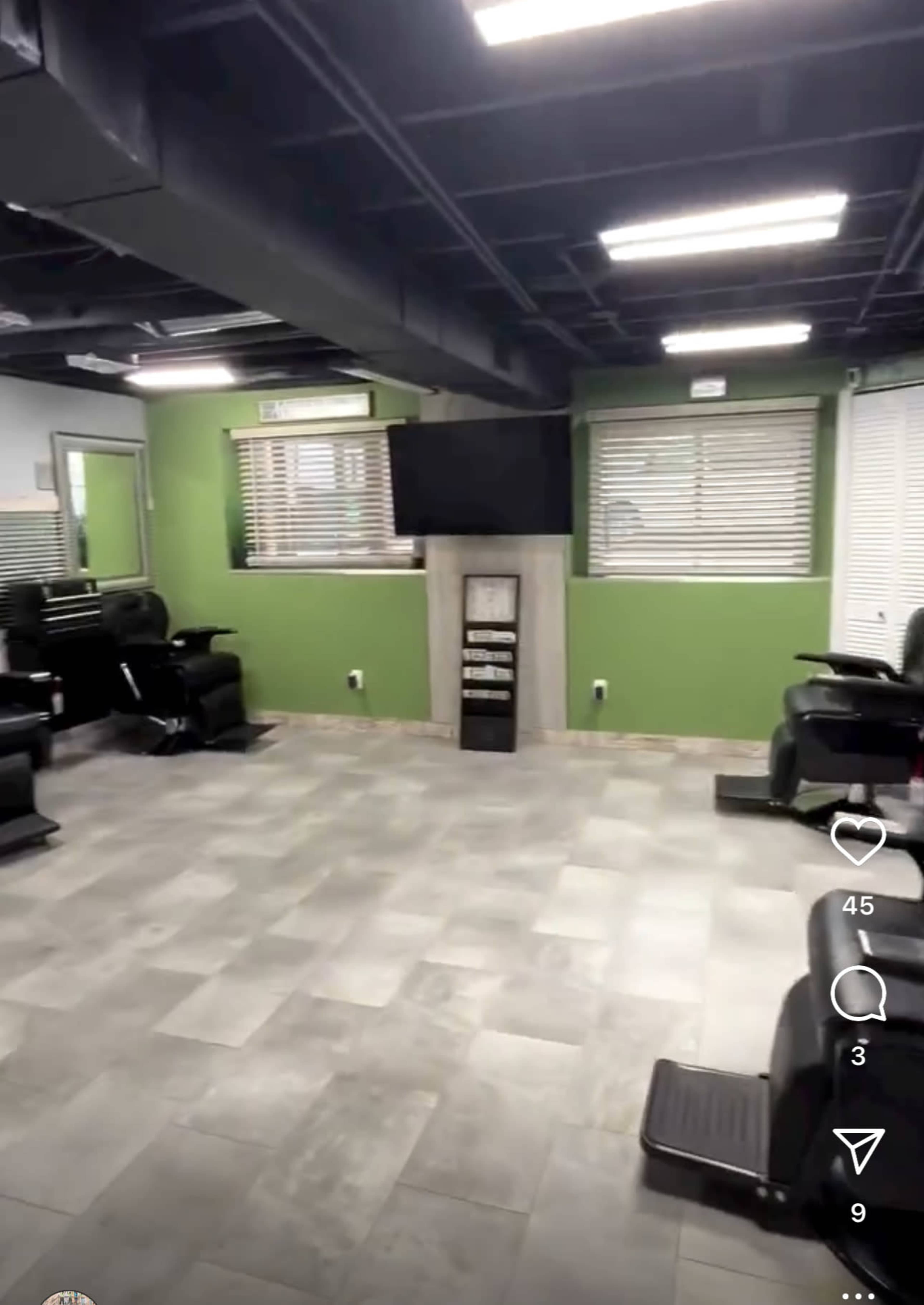 The interior of a salon featuring several black salon chairs arranged around a central open space, with a large TV mounted on a wall and light green walls.