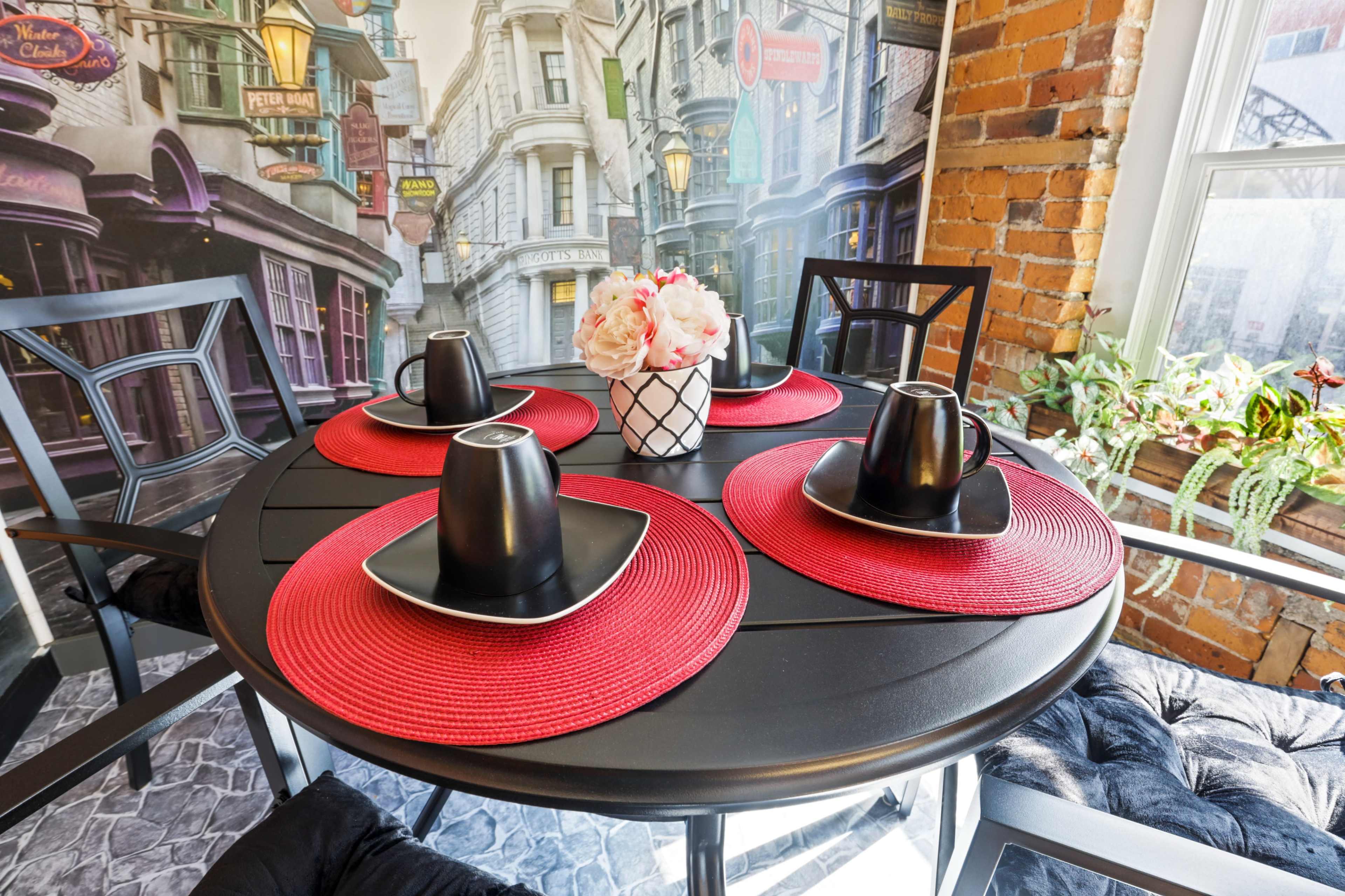 A circular black dining table is set with four black dishes and cups on red placemats, surrounded by brick walls and a wallpaper depicting a street scene.