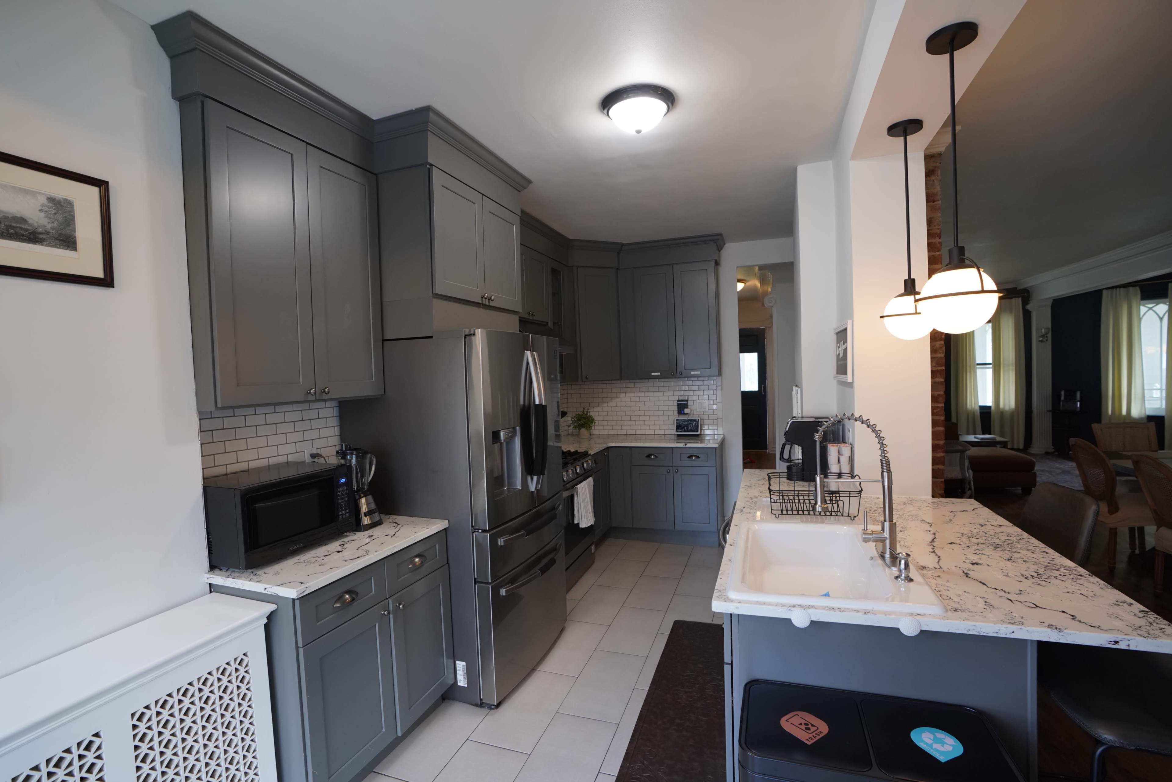 The image shows a modern kitchen with gray cabinets, a stainless steel refrigerator, and a white farmhouse sink next to a dark countertop.