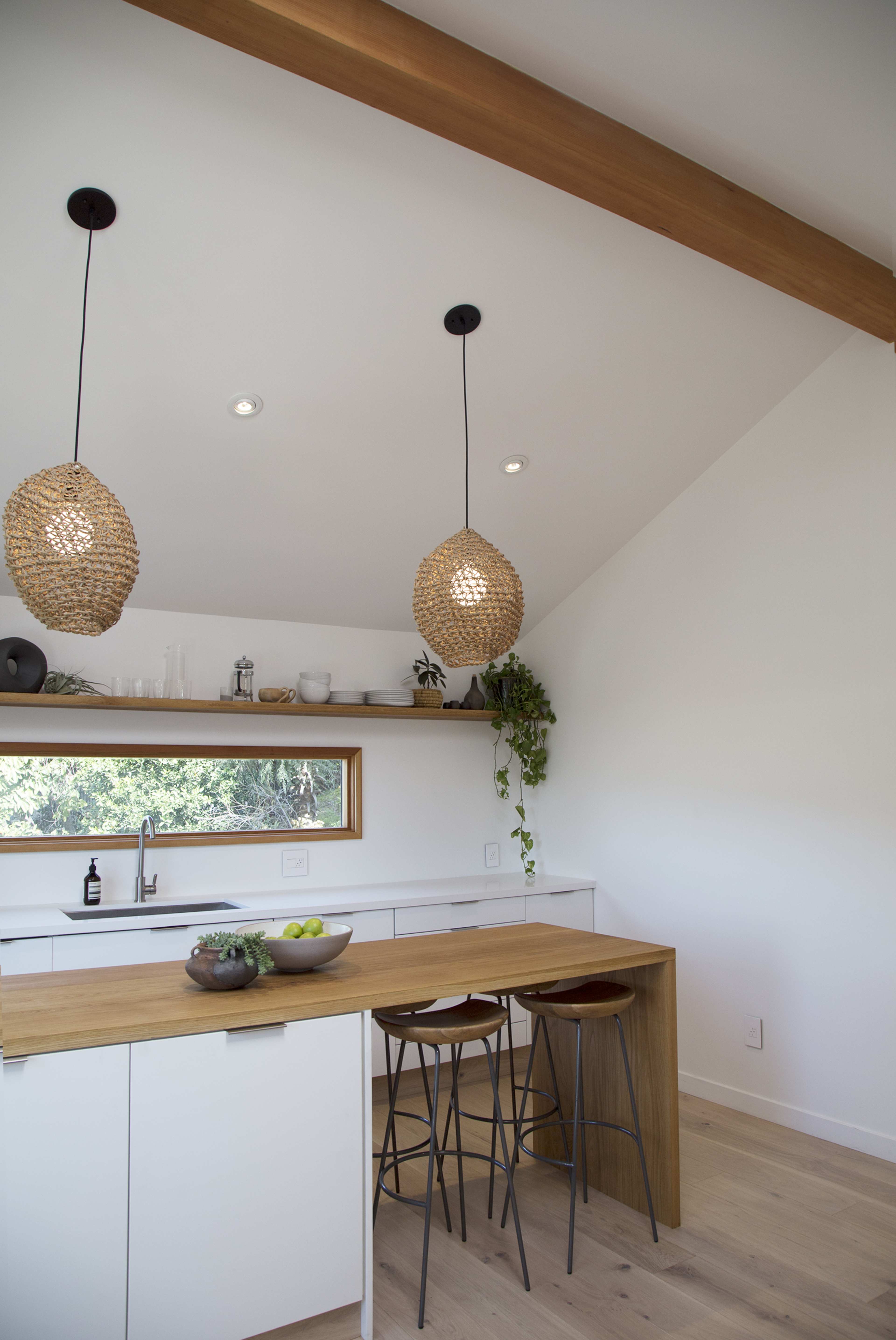 A bright kitchen features a wooden countertop, sleek cabinetry, and hanging woven light fixtures above two metal stools.