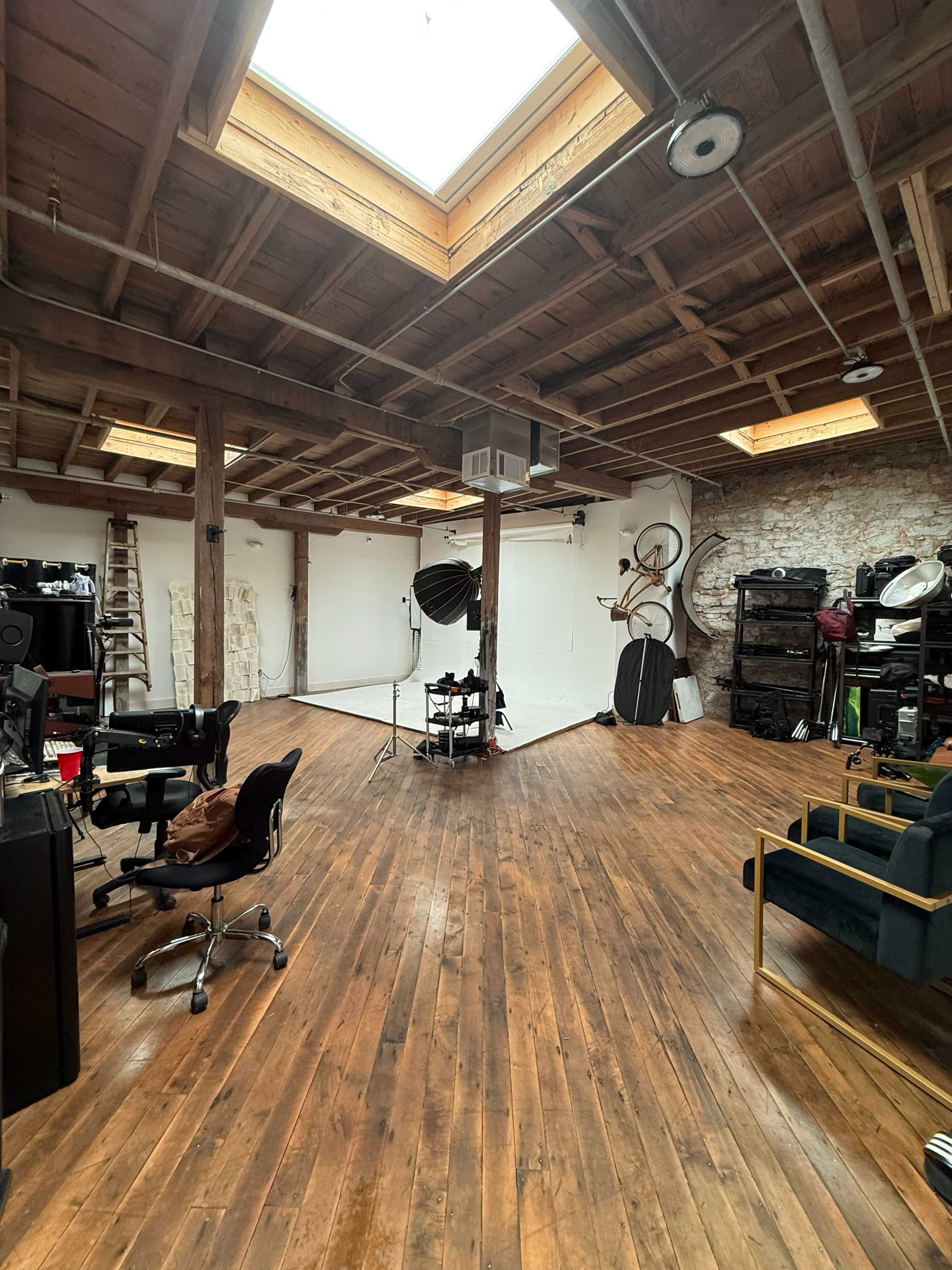 The image shows a creative studio space with wooden floors, skylights, and various photography equipment arranged around a central area featuring a white backdrop.