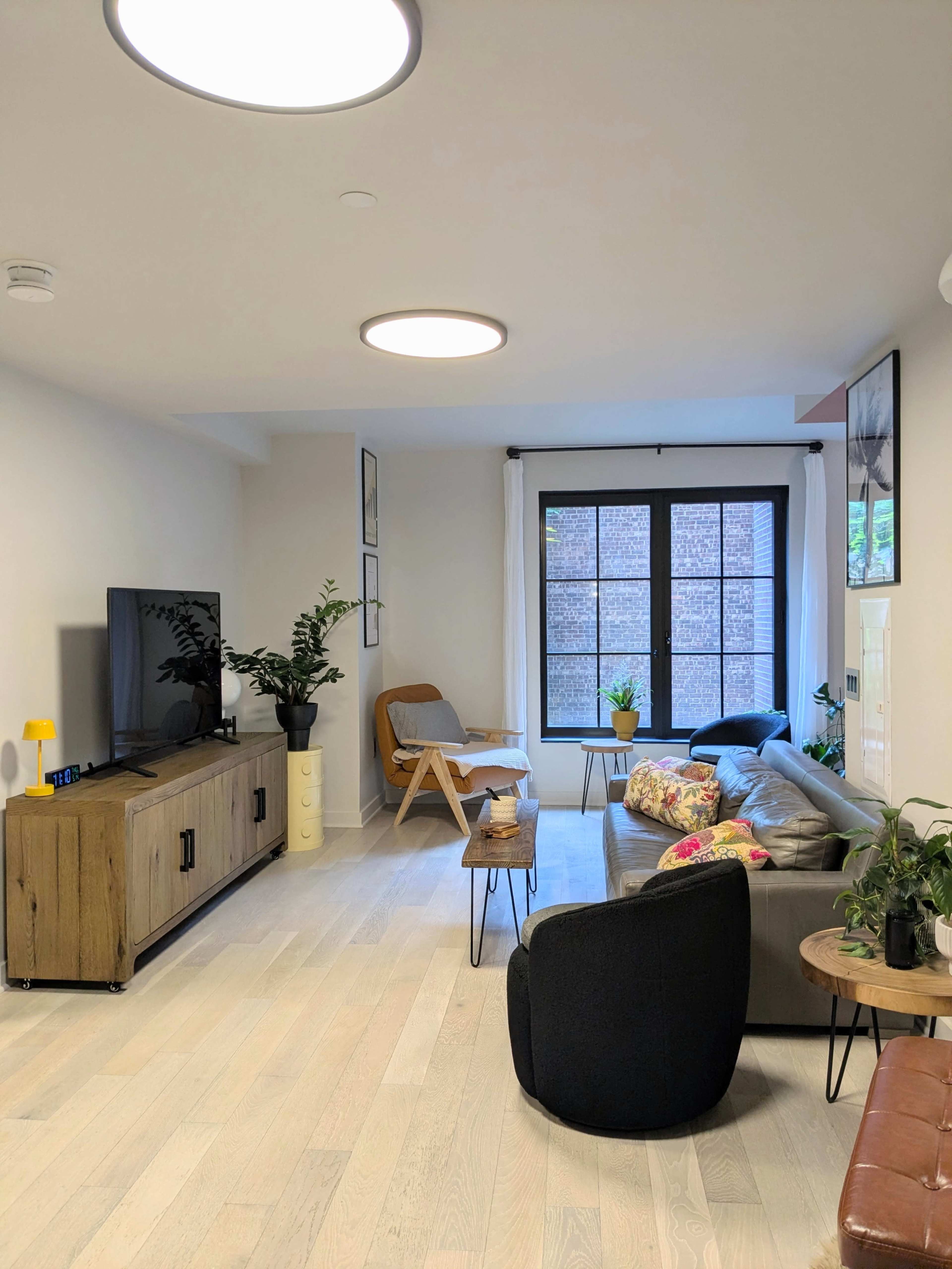 The image shows a modern living room featuring a television unit, a sofa with decorative pillows, a chair, and several potted plants near a large window.