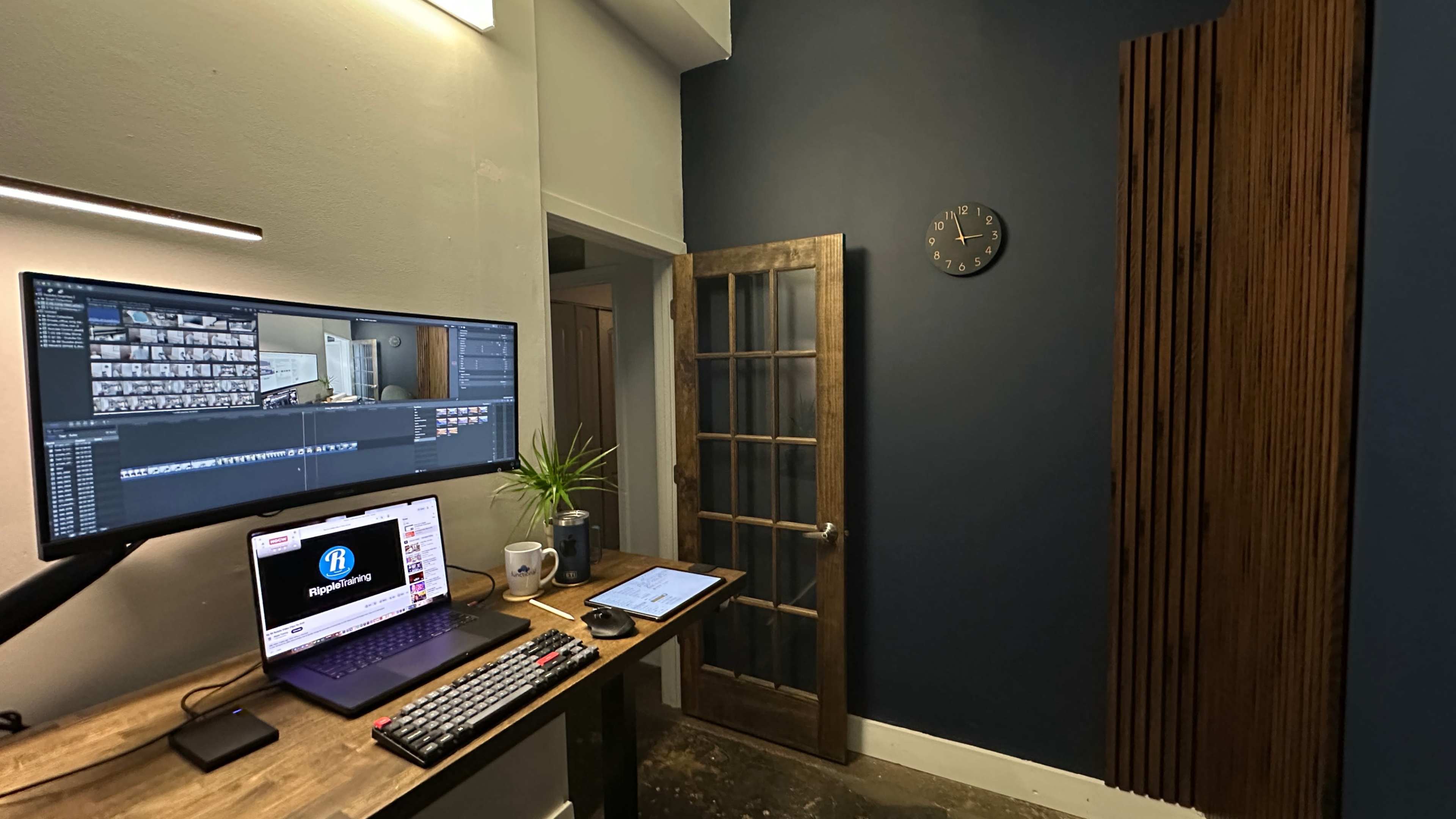 A workspace features a computer monitor and laptop on a wooden desk, with a clock on the wall and a door leading to another room.