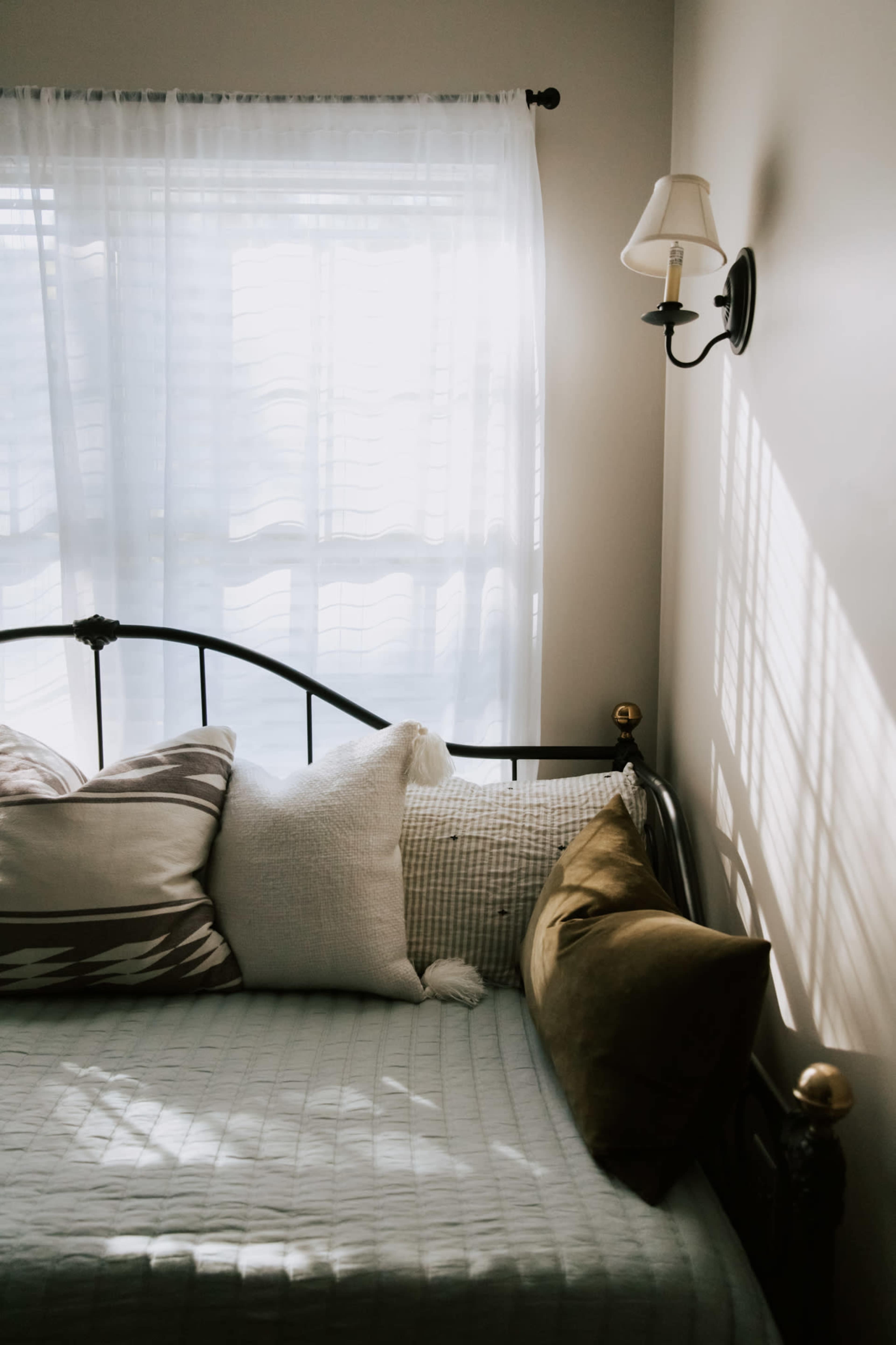 A bed with several decorative pillows is positioned beside a window, which has sheer curtains allowing light to filter through.
