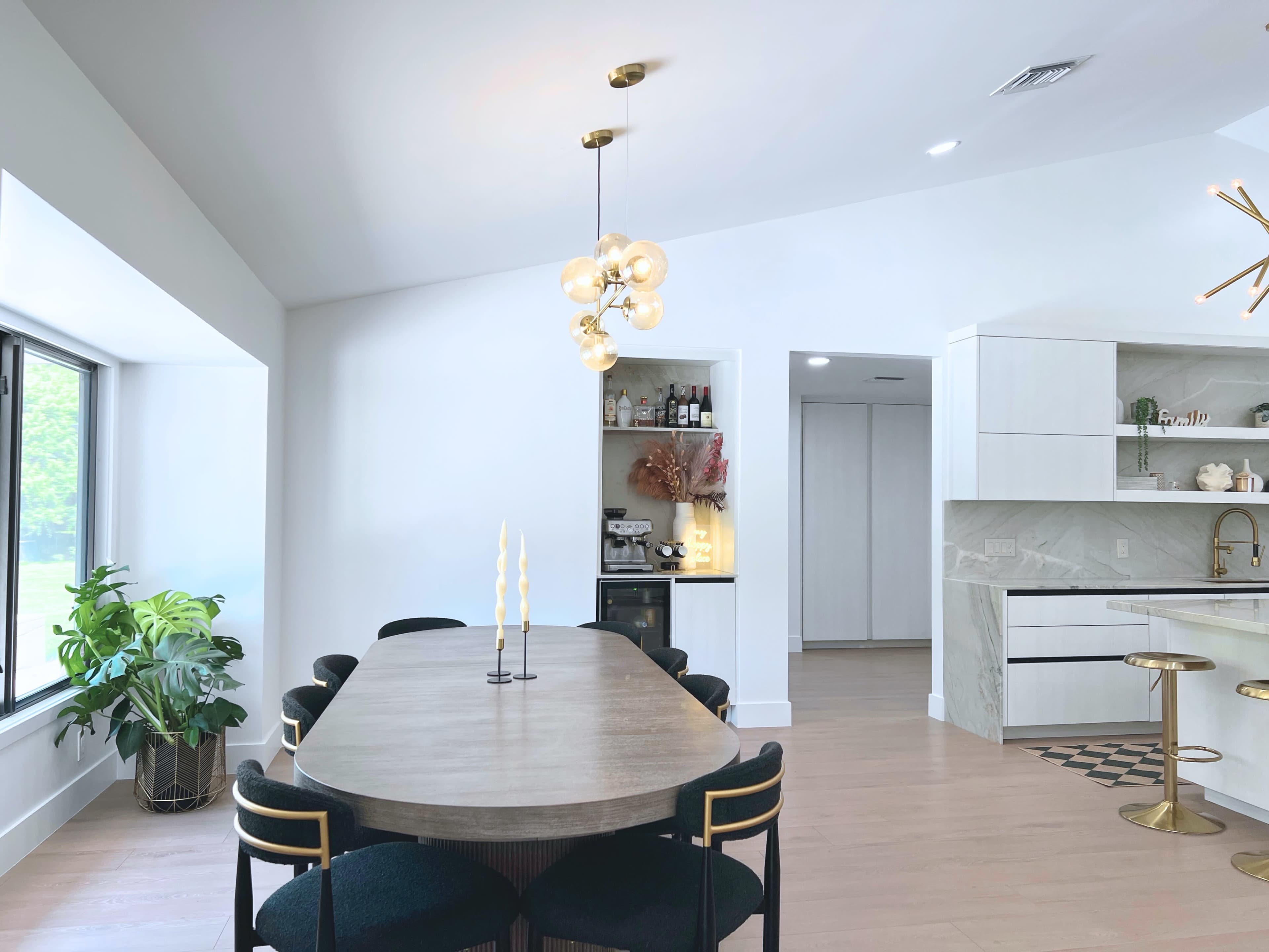 A modern kitchen and dining area features a large wooden table, sleek black chairs, a pendant light, and a stylish kitchen with marble accents and a bar counter.
