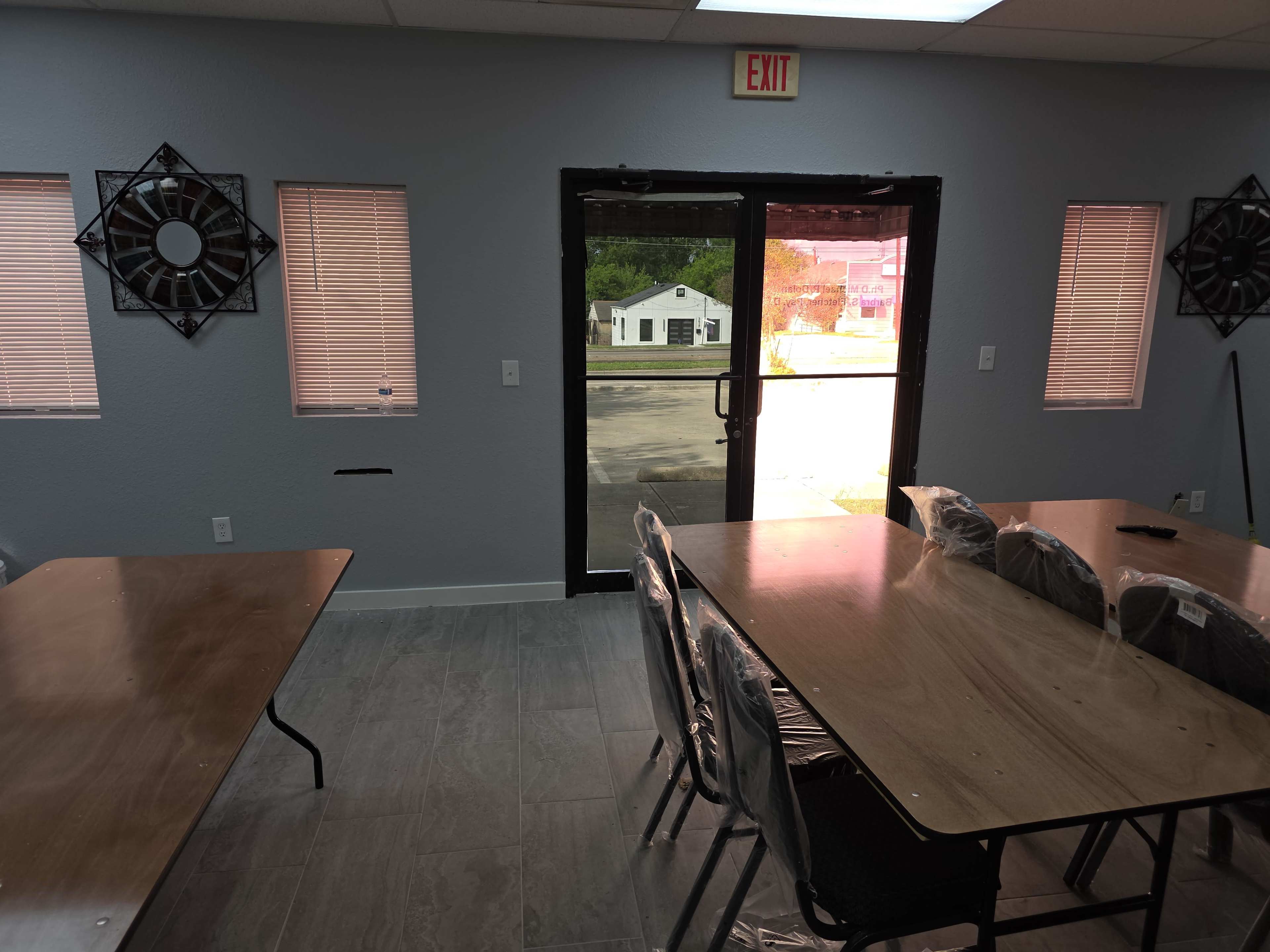 The image shows a partially furnished room with several wooden tables and black chairs, and a doorway leading to an exterior view.