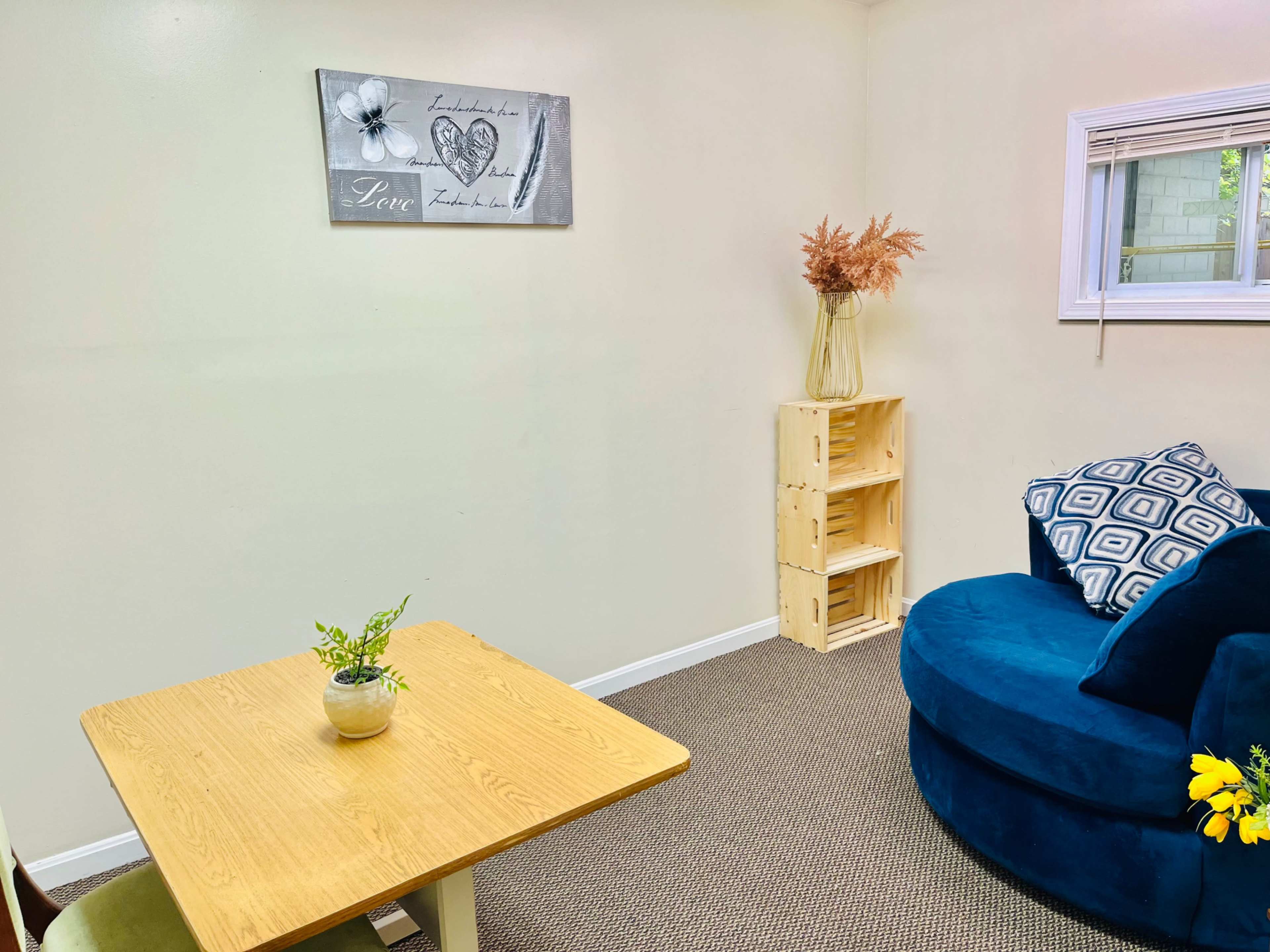 A small, well-lit room contains a blue circular sofa, a wooden table with a plant, and a shelving unit in the corner.