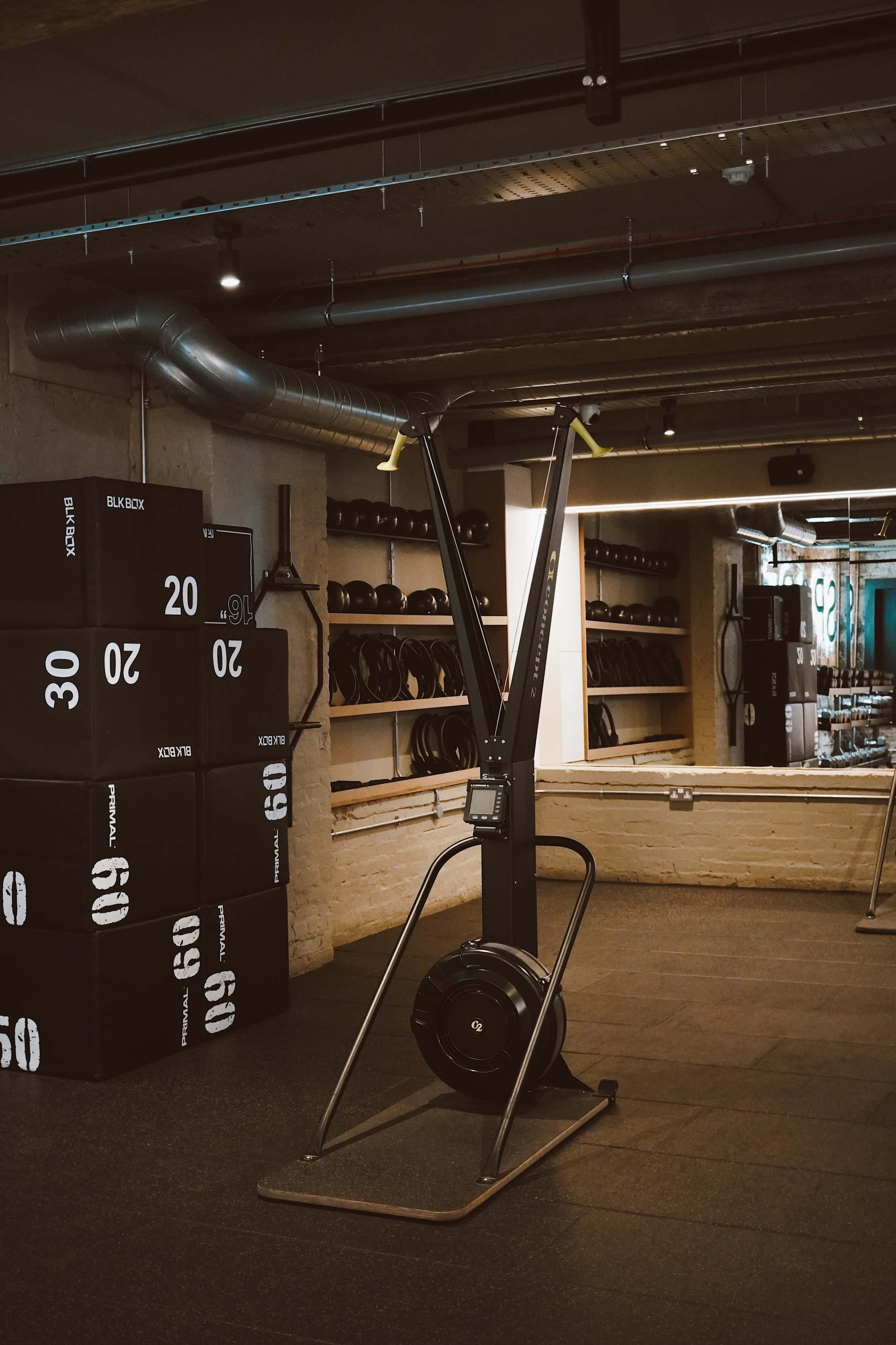 A rowing machine is positioned in a gym with stacked black weights and a mirror in the background.