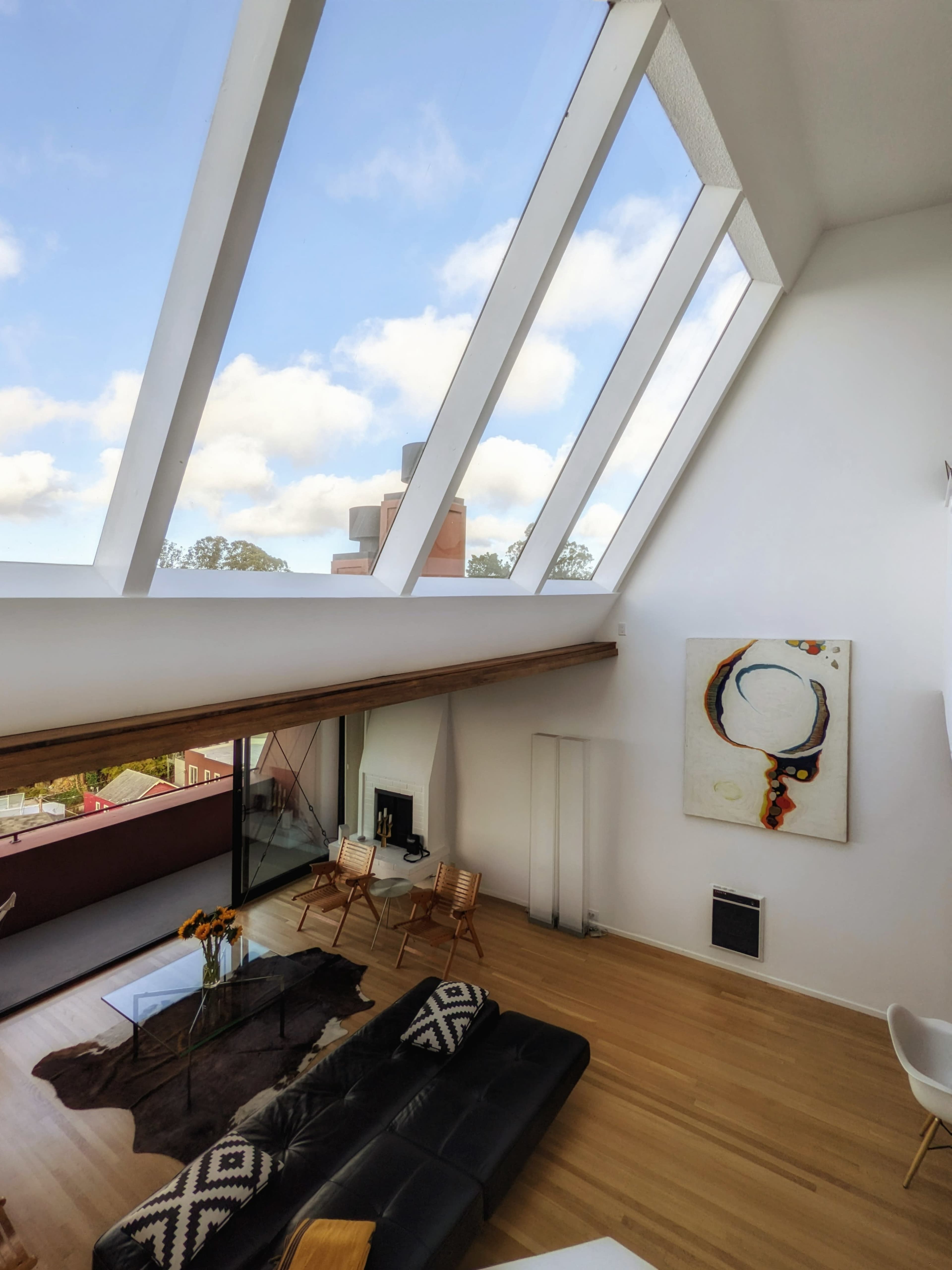 The image shows a spacious, modern living room with large skylights, a black sofa, a glass coffee table, and a colorful piece of art on the wall.