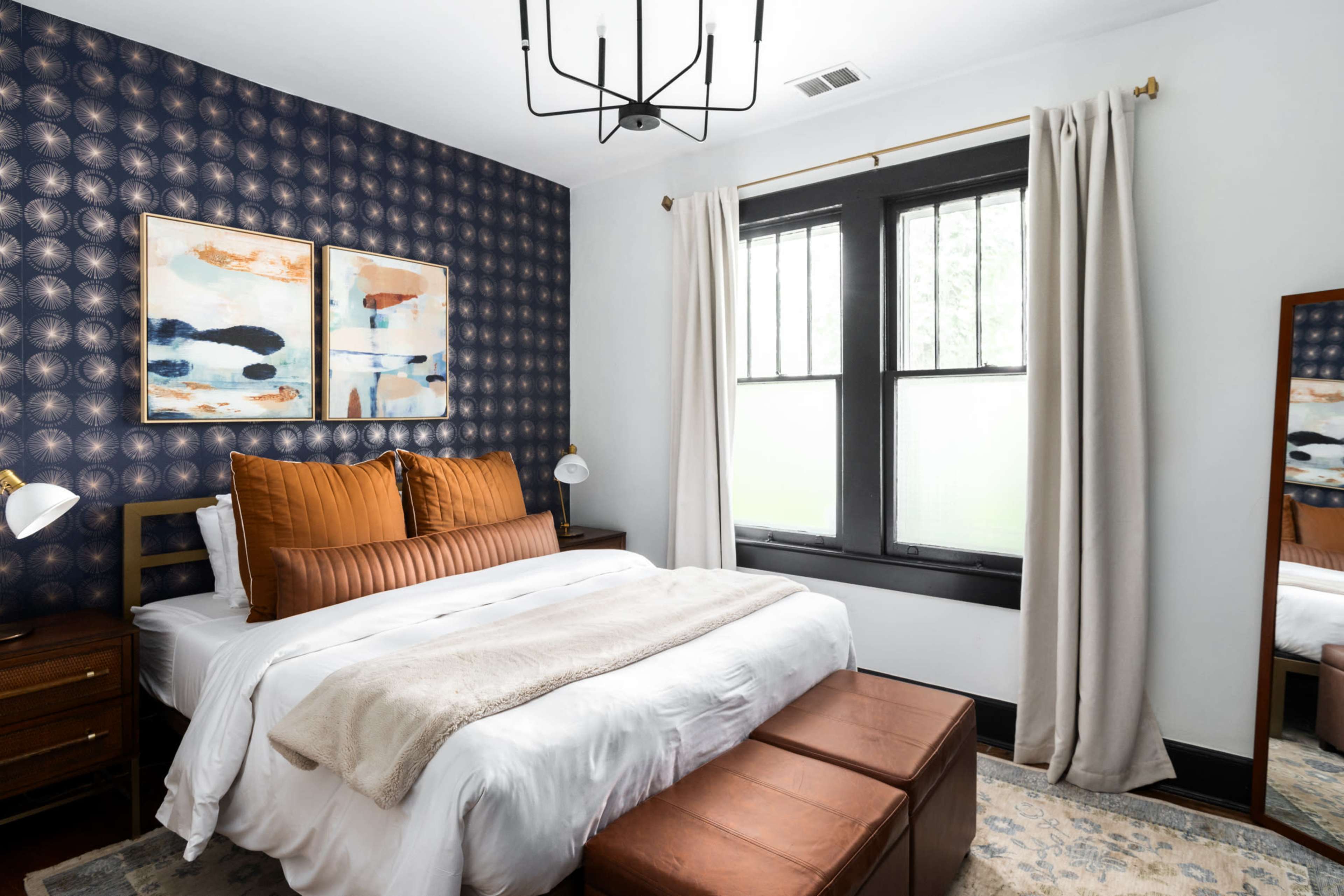 A cozy bedroom features a patterned accent wall, a bed with decorative pillows, and a large mirror beside a window.