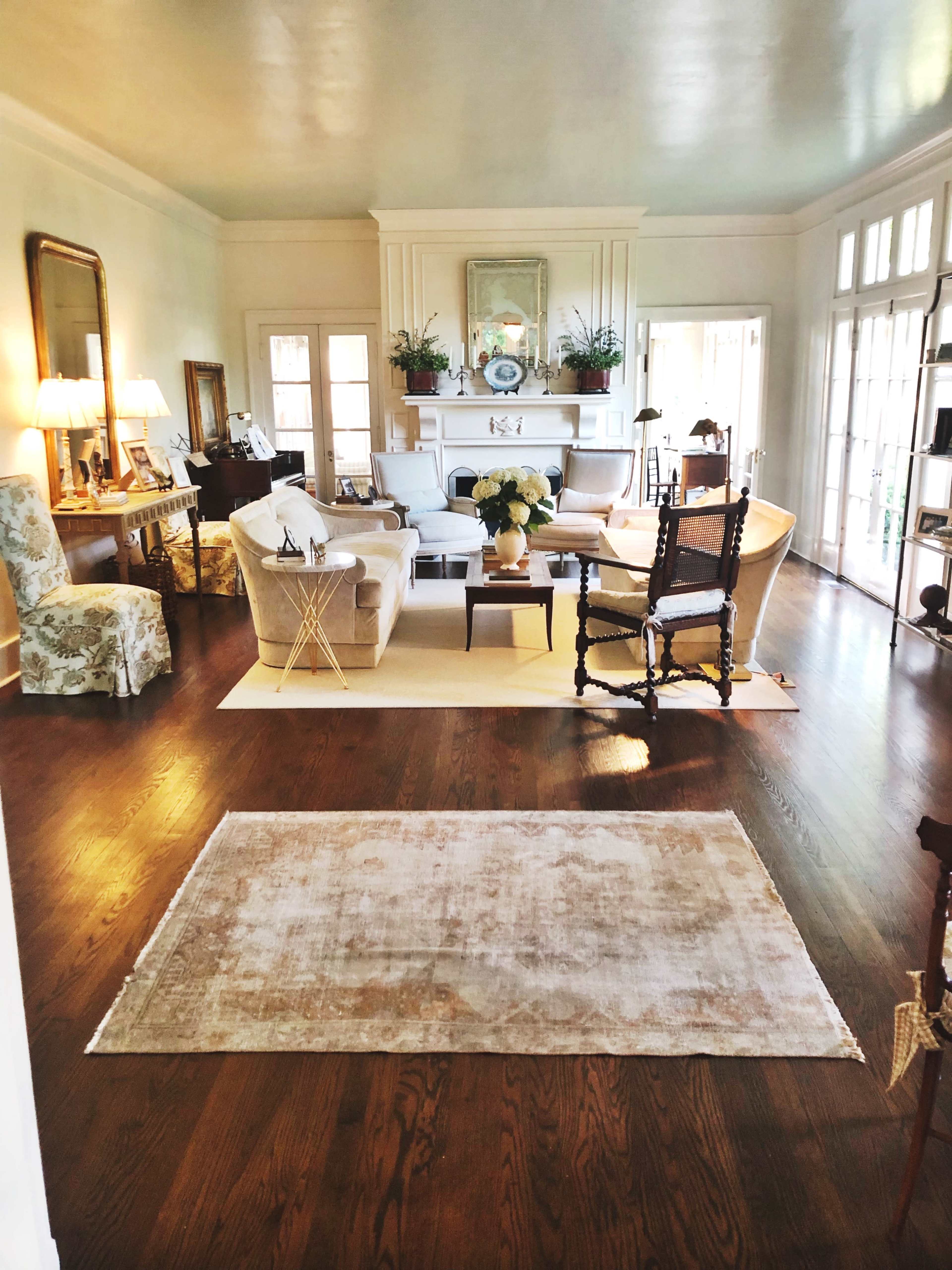 A spacious living room features a central seating area with a sofa, armchairs, and a coffee table, surrounded by large windows and decorative plants.