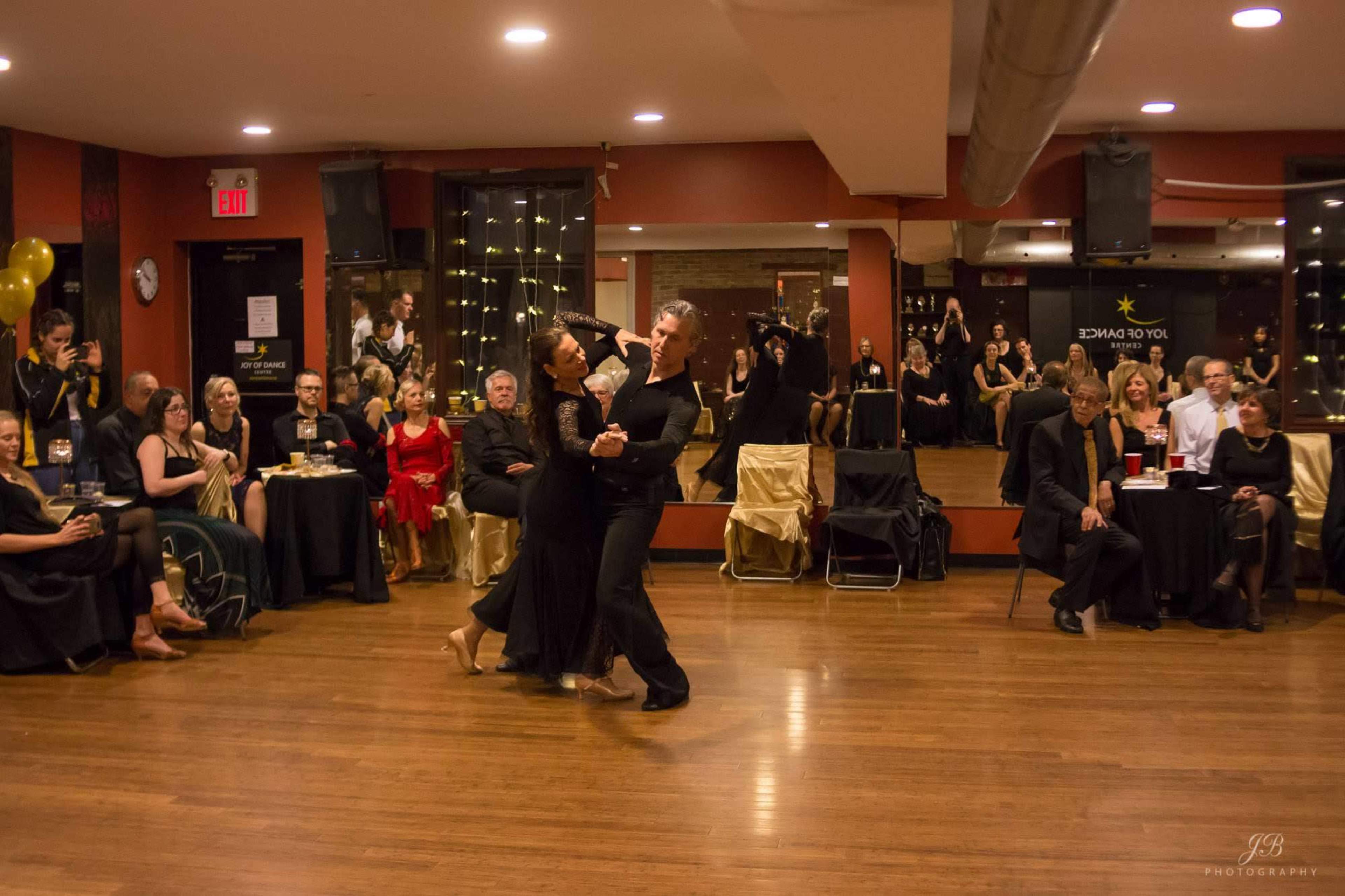 A couple dances in formal attire on a polished wooden floor as an audience watches from seated tables in a decorated event space.