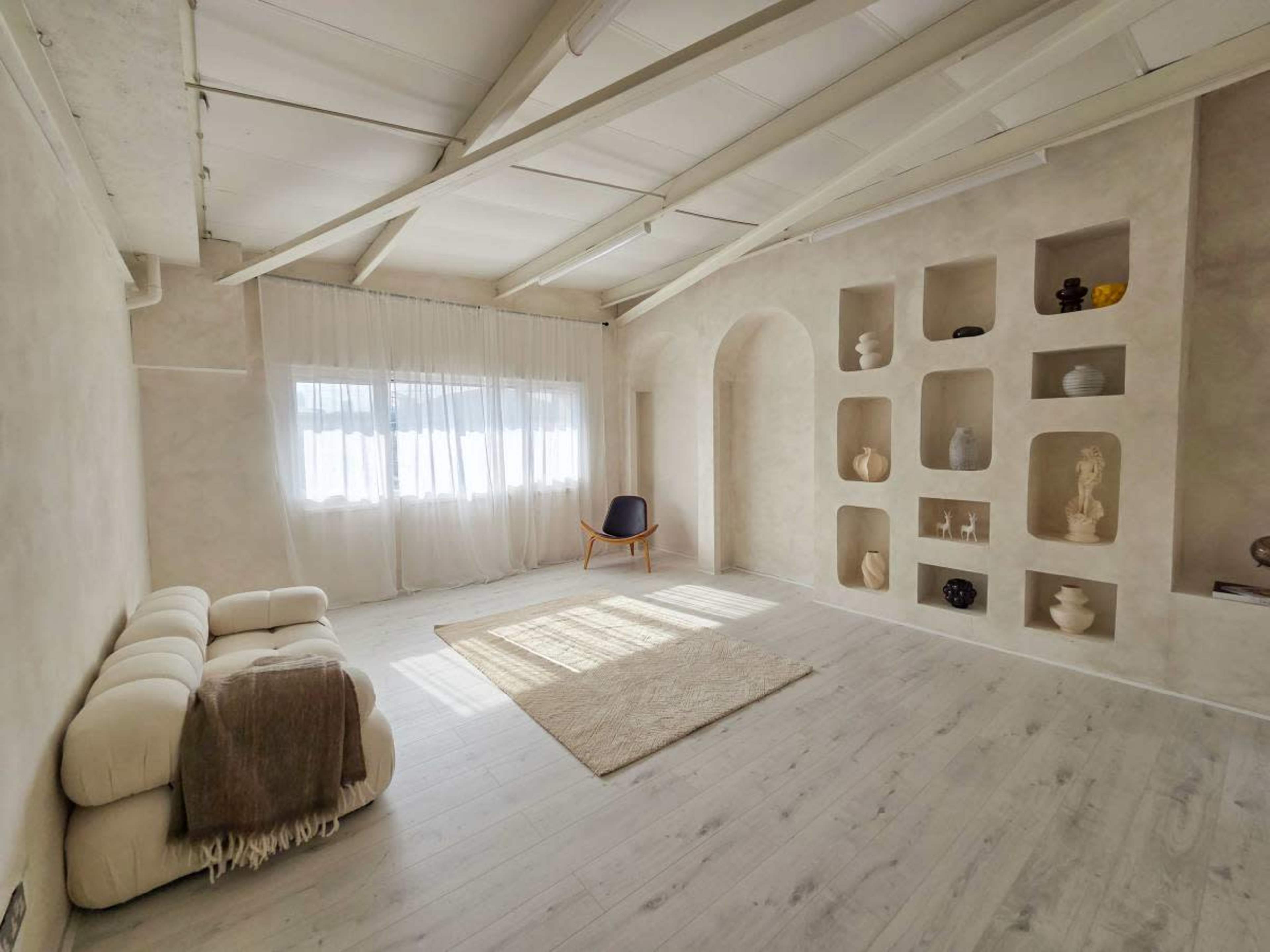 The image shows a minimalist interior with light-colored walls, wooden beams, a large window with sheer curtains, and a decorative shelving unit displaying various ceramic objects.